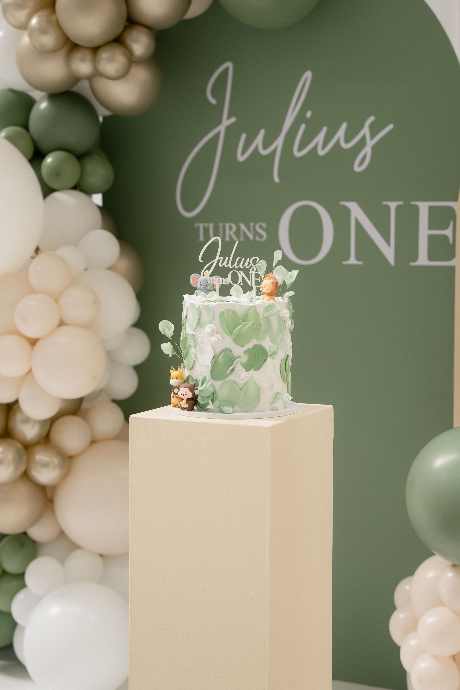 Green and cream balloon arch frames a first birthday cake with Julius Turns One topper on a beige pedestal.