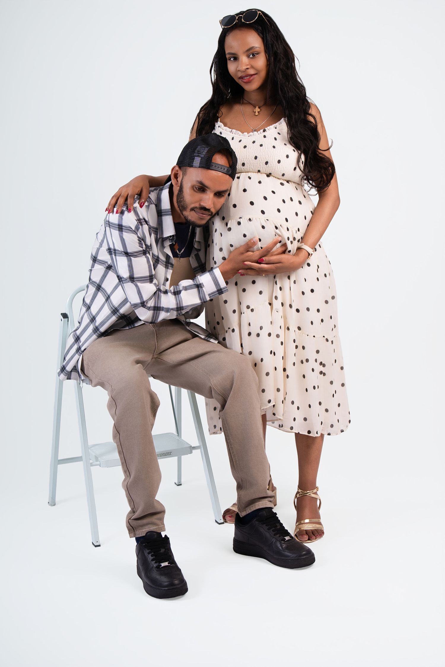 A man sitting on a stool leans toward a pregnant woman in a polka dot dress, both posing for a maternity photo.