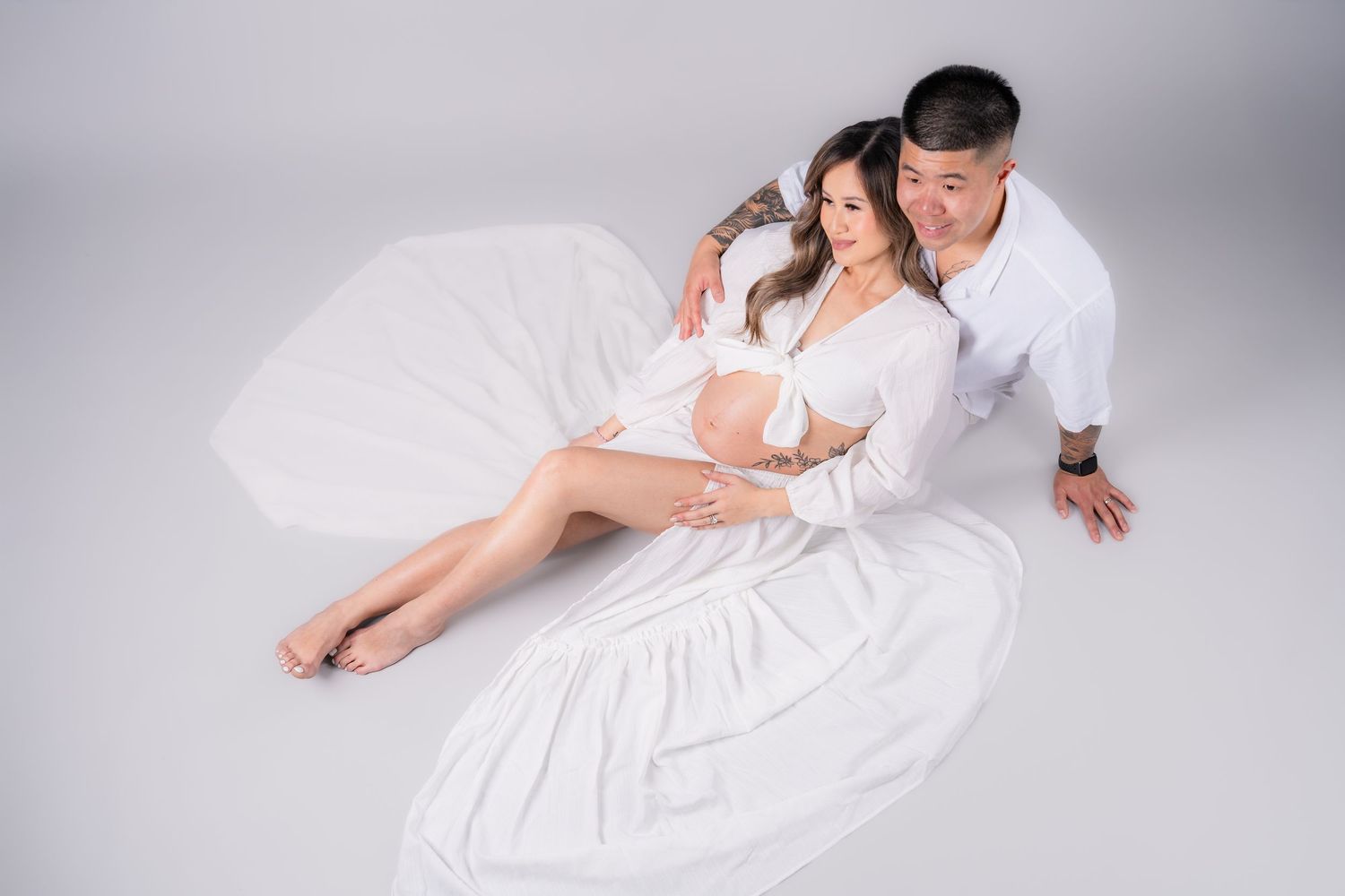 Pregnant couple in white outfits posing on studio floor, woman in flowing skirt with man embracing her baby bump.