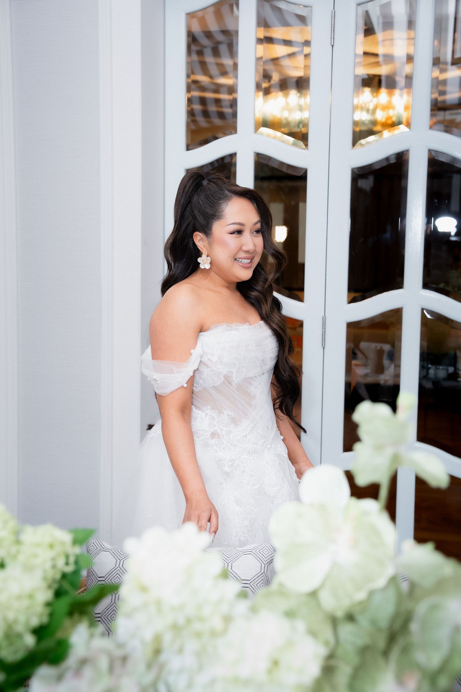 Bride in off-shoulder lace wedding gown smiling near elegant white French doors with floral arrangements in foreground.