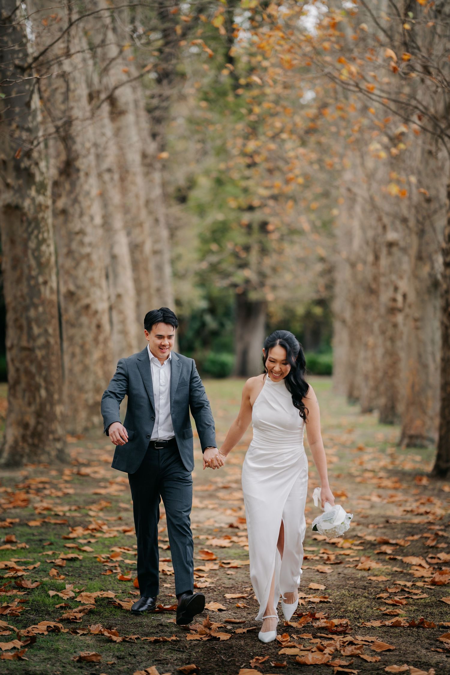 Couple walks hand-in-hand through autumn tree-lined path, bride in white dress, groom in gray suit, fall leaves on ground.