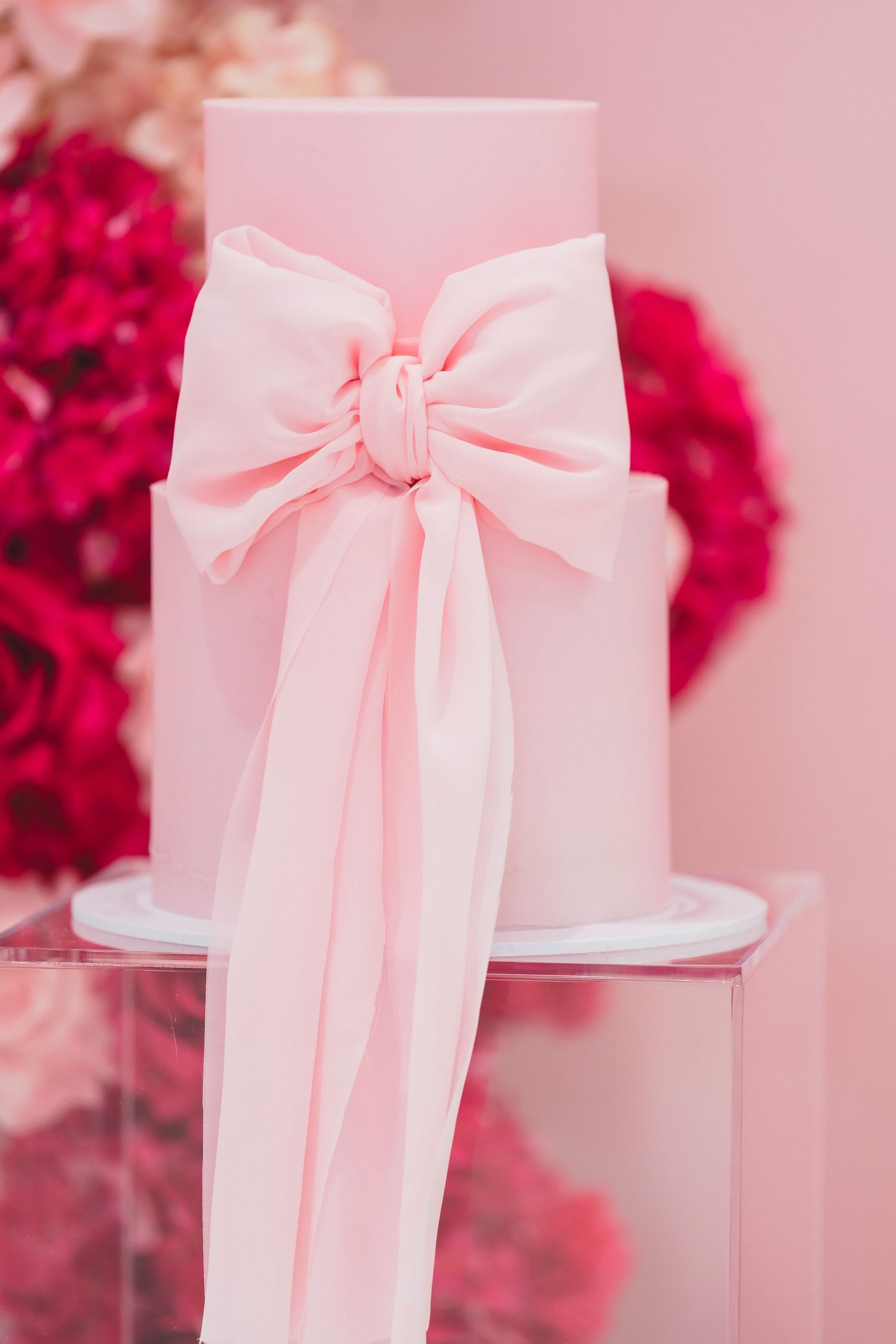 Elegant two-tier pink wedding cake decorated with oversized satin bow on pink floral background.