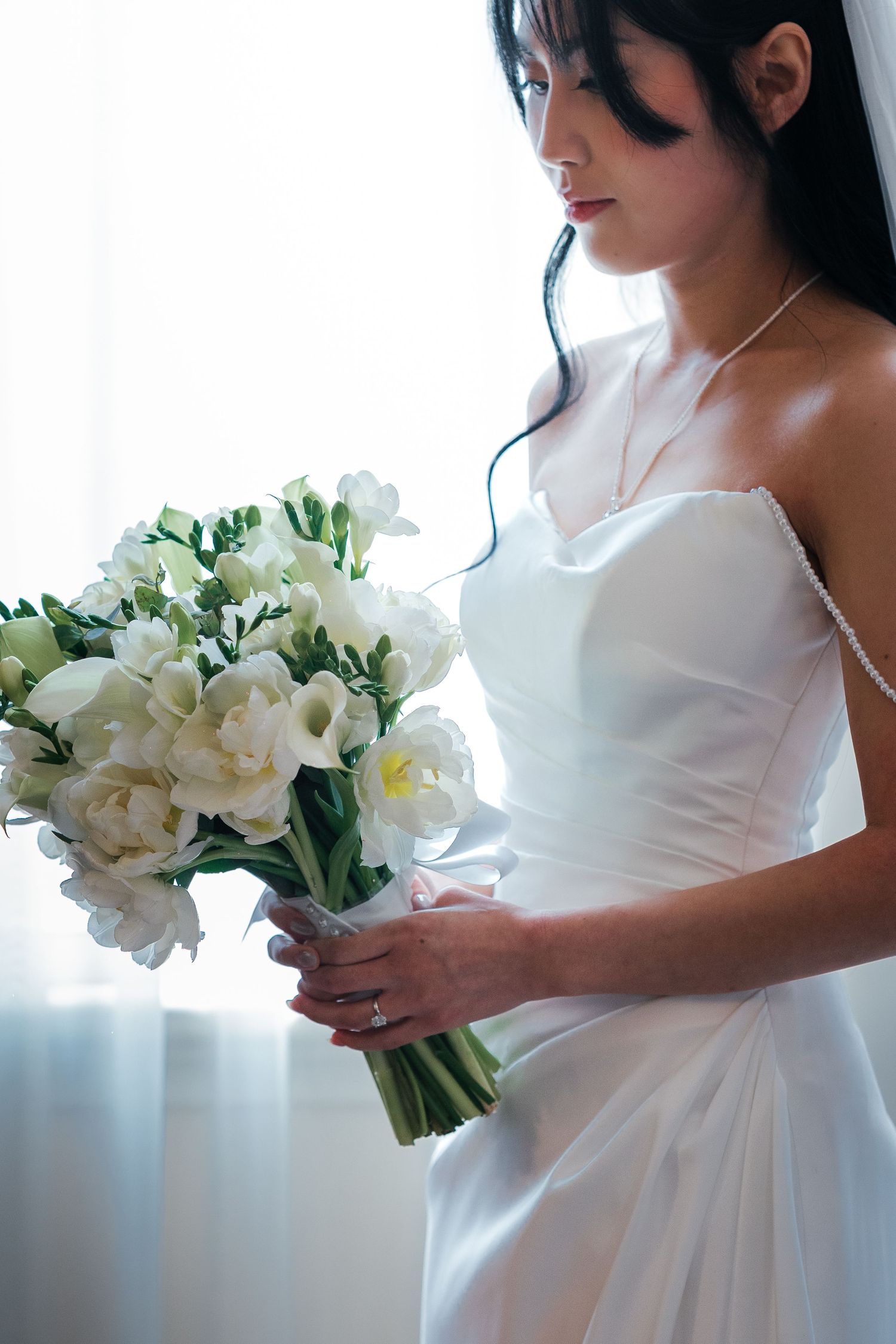 Bride in white strapless gown holding elegant bouquet of white flowers, looking down gracefully.