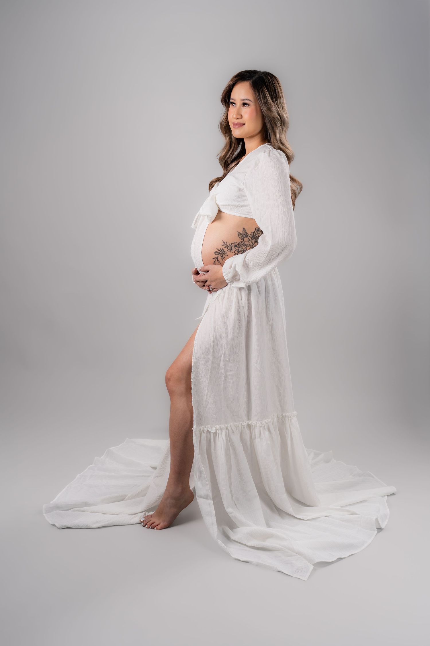 Pregnant woman in white flowing maternity gown with tattoos cradling baby bump in professional studio photo.