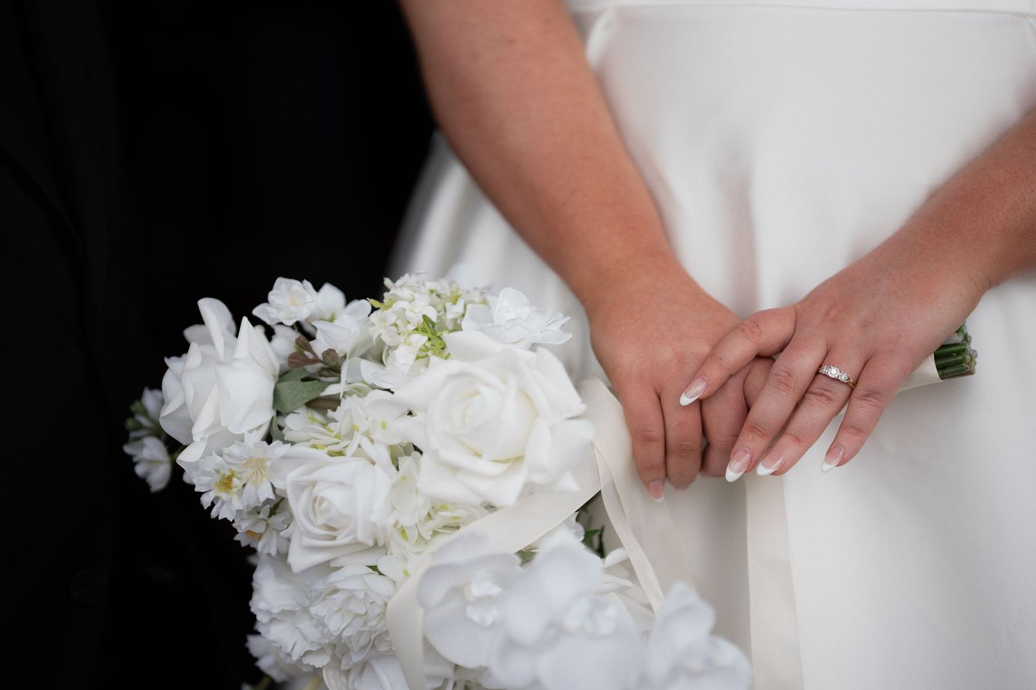 Bride holding white rose bouquet with wedding ring visible on hand, wearing white dress beside groom in black suit.