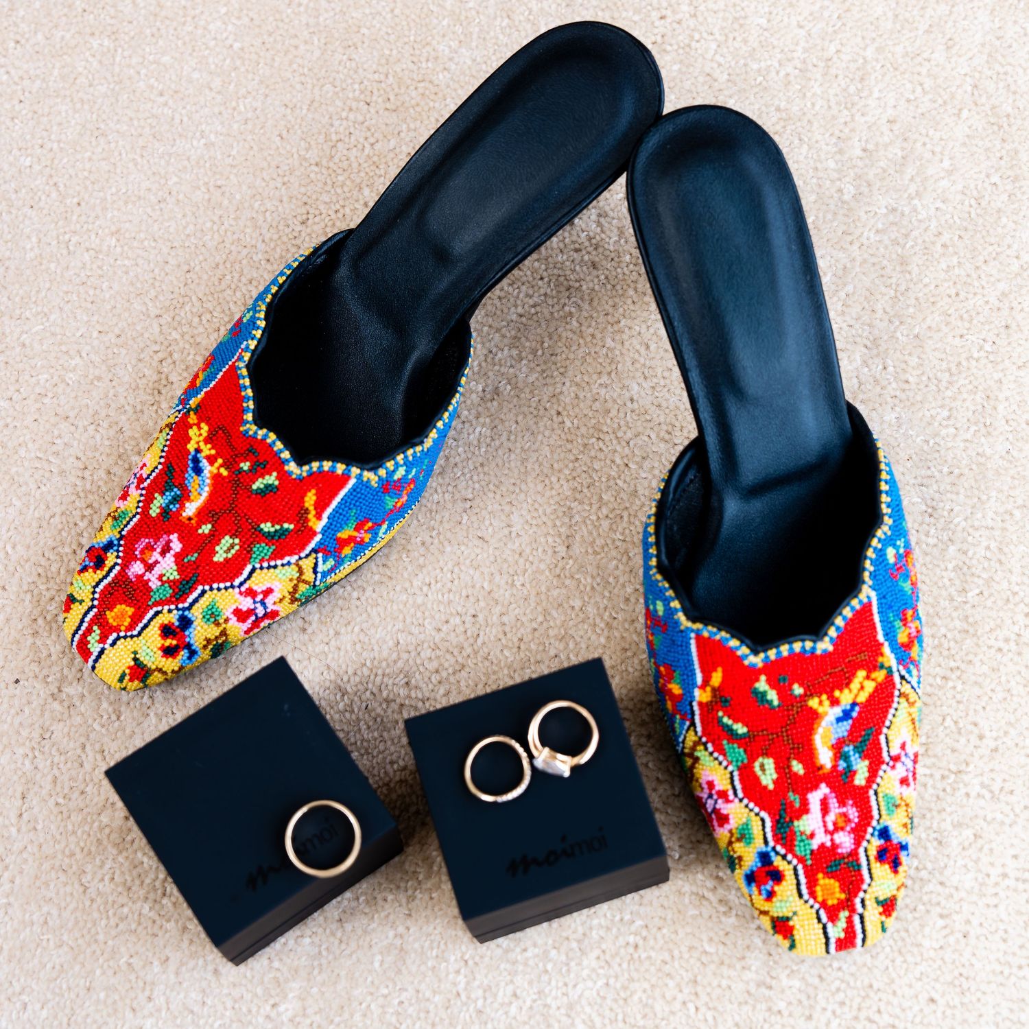 Colorful embroidered floral mule slippers with beaded trim displayed beside two gold rings on black jewelry boxes.