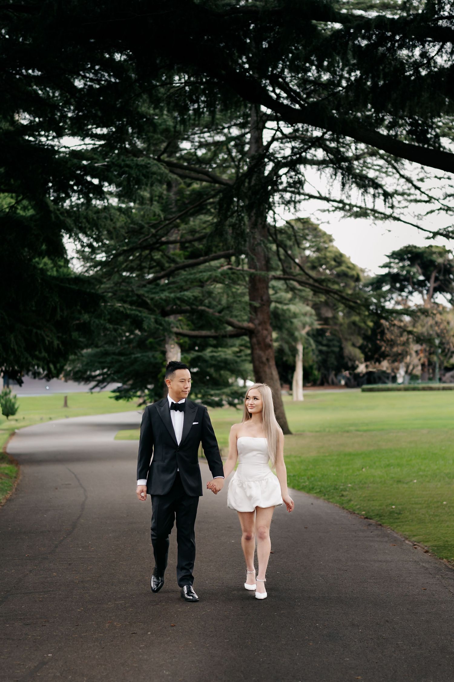 A couple in formal attire walks hand in hand down a tree-lined path.