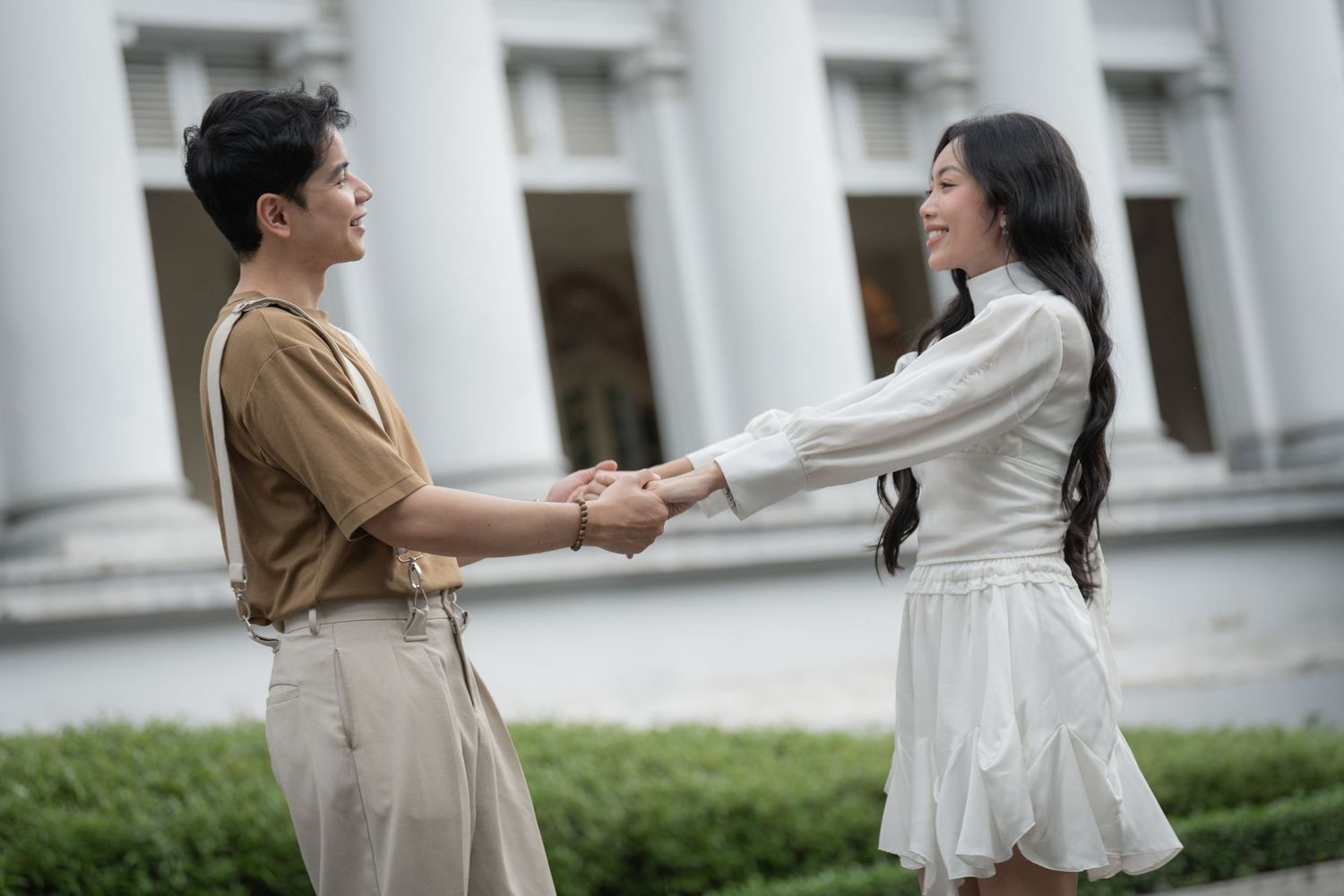 Couple in casual attire poses playfully by white pillars outdoors.