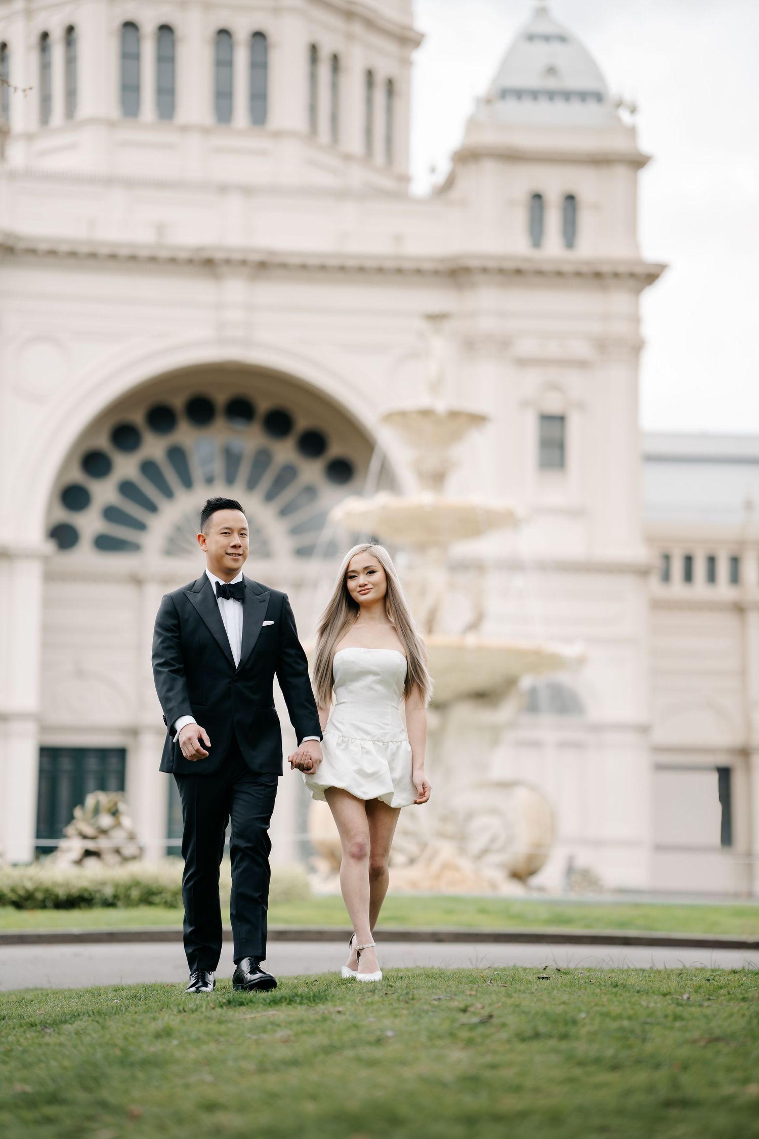 A couple walks together on grass in front of an ornate building with classical architecture.