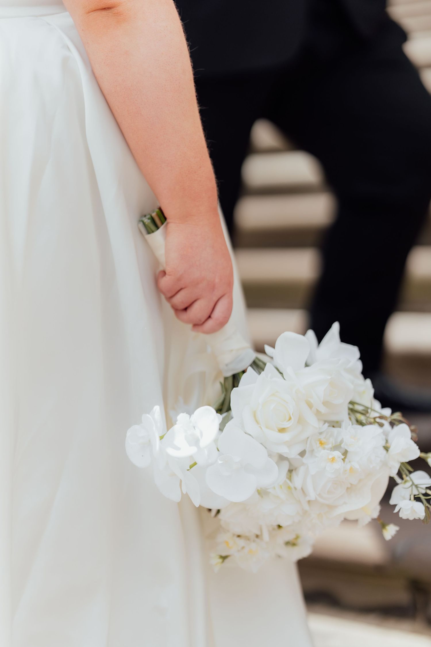 Bride holding a white wedding bouquet of roses and orchids in her right hand, wearing a flowing white gown.