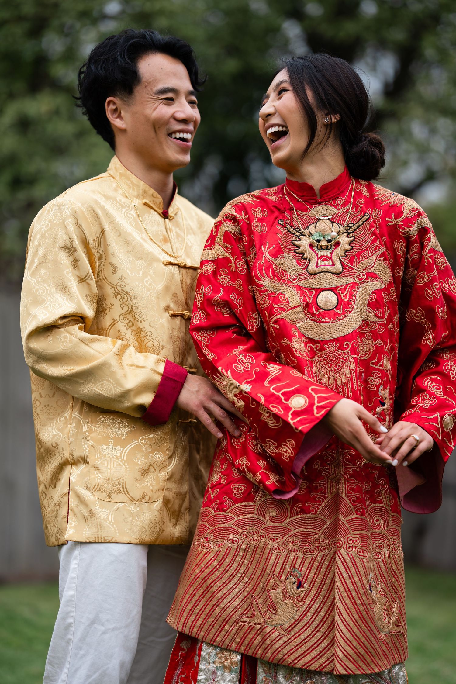 A smiling couple in traditional Chinese wedding attire, man in gold brocade and woman in red dragon embroidered jacket.