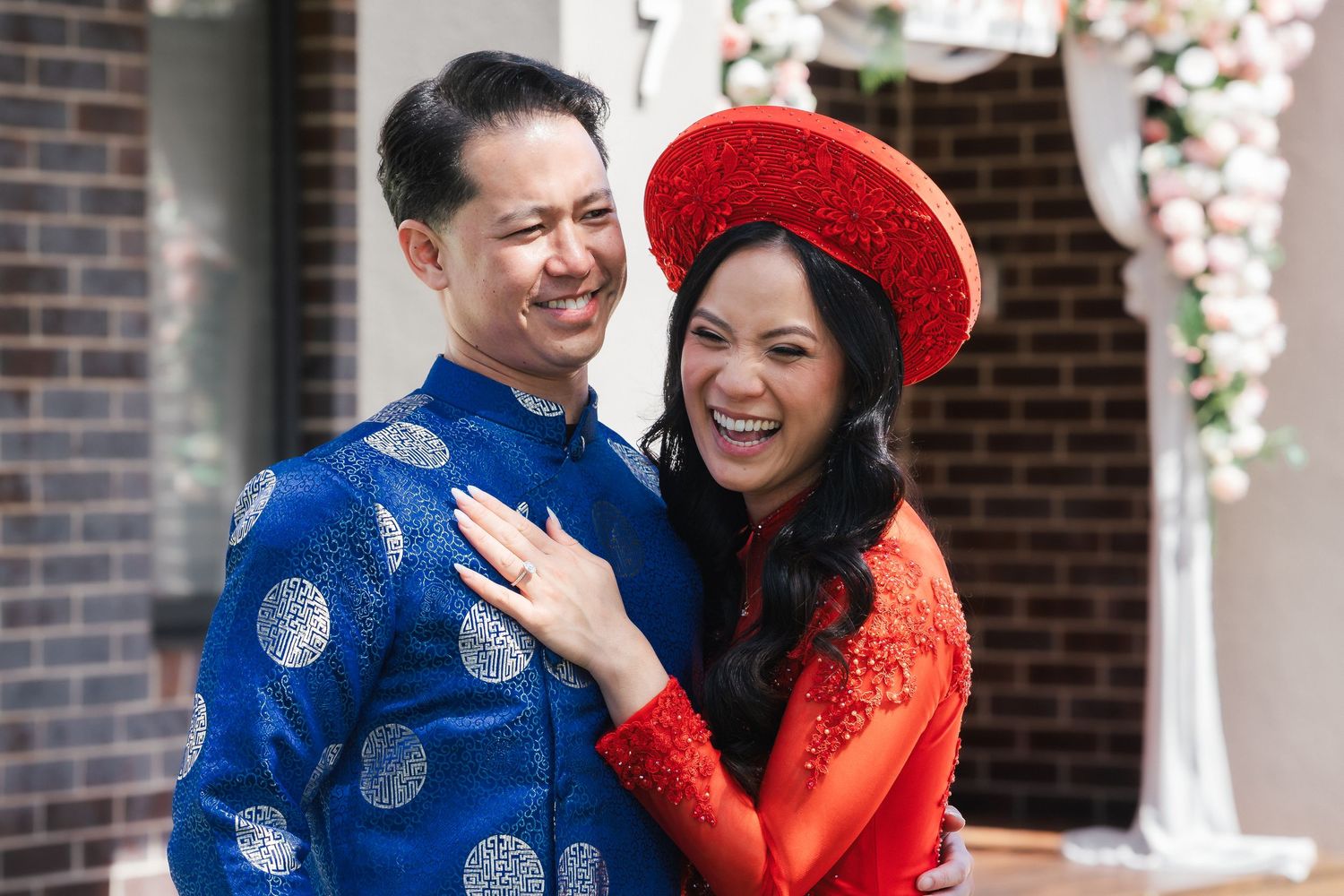 Happy couple in blue and red Vietnamese ao dai traditional wedding attire embracing outdoors.