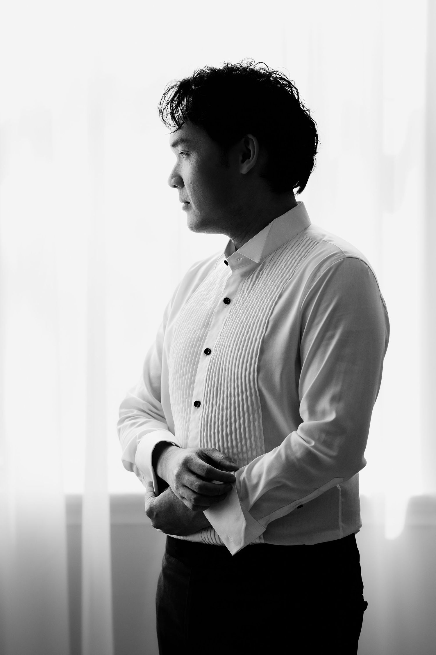 Black and white portrait of a groom in a white dress shirt buttoning his cuff by a bright window.