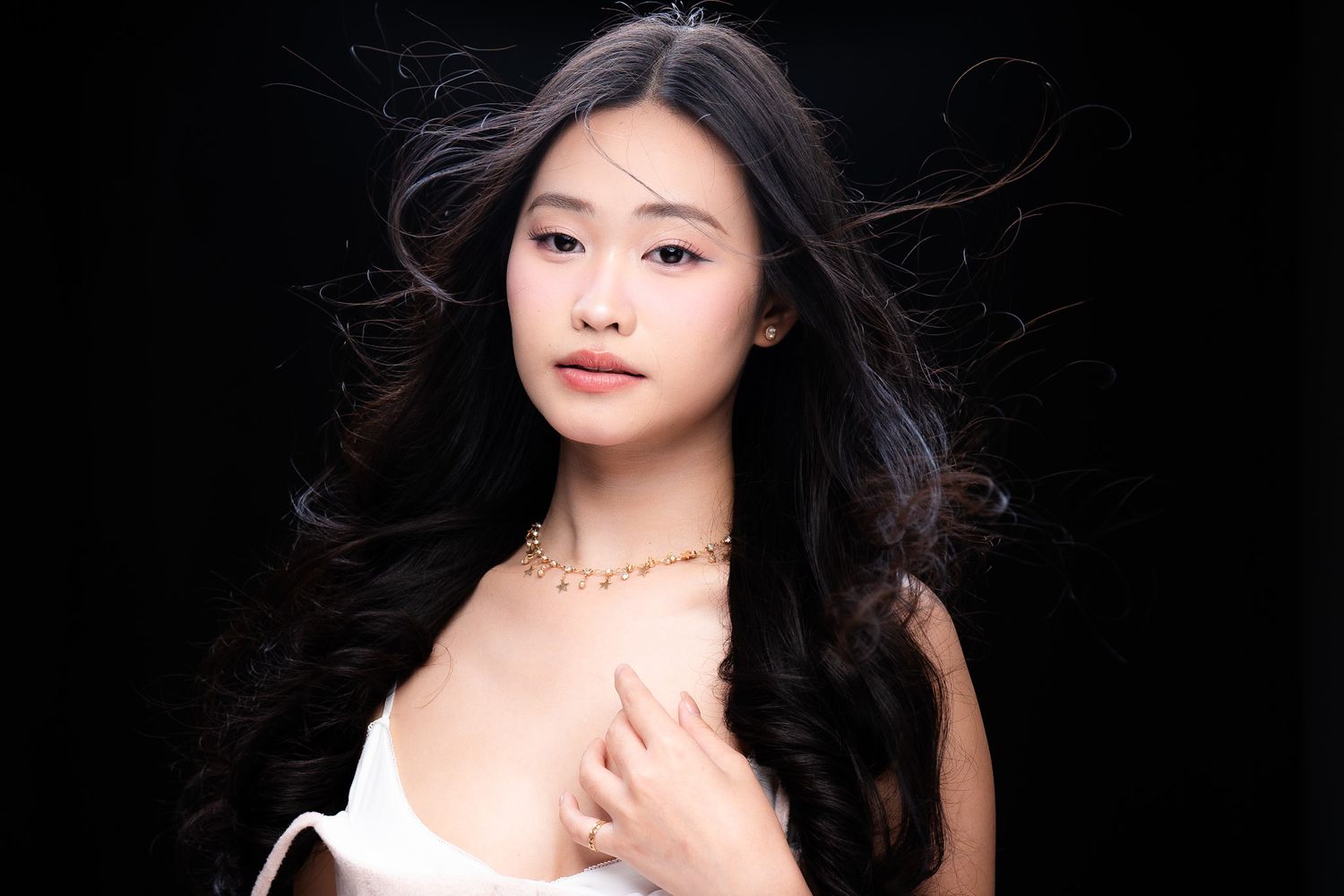 Young Asian woman with long wavy black hair wearing a gold necklace and white top against dark background.