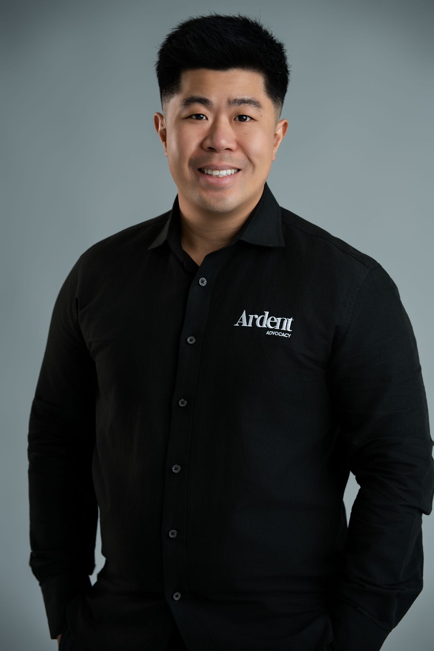 Professional man in black Ardent Advisory branded shirt smiling confidently against gray background.