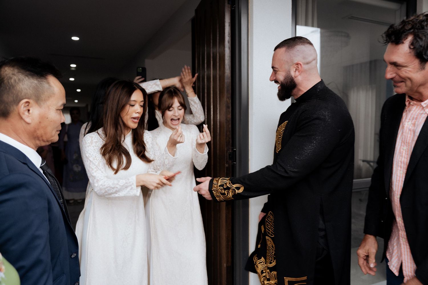 Guests in white ao dai greet man in black suit at Vietnamese wedding ceremony.