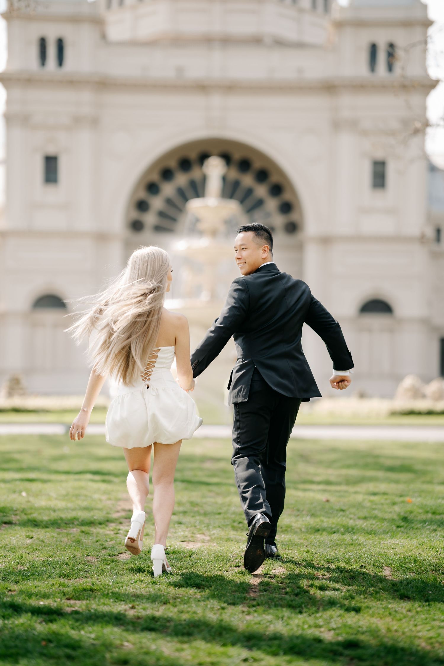 A couple in formal attire playfully runs across a grassy lawn near a grand building.