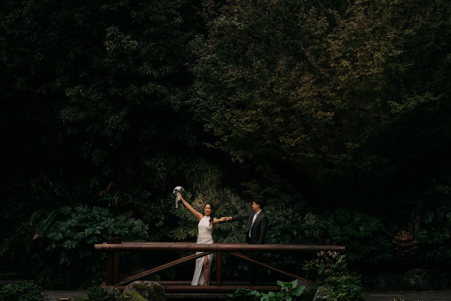 A bride and groom celebrate on a wooden bridge surrounded by lush green forest trees on their wedding day.