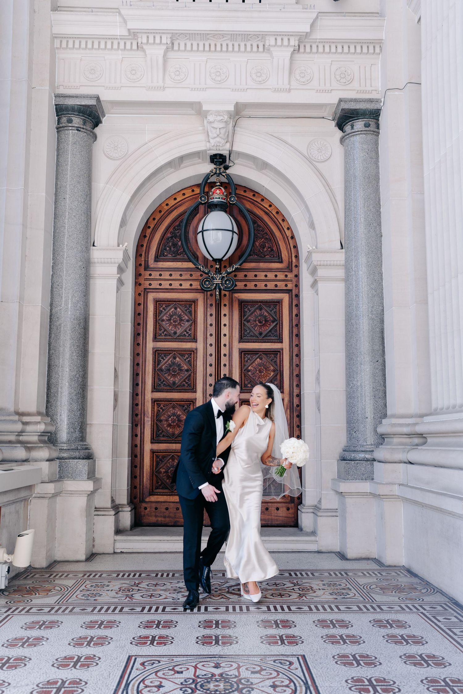 Elegant couple posing in front of ornate white building with arched wooden doors.