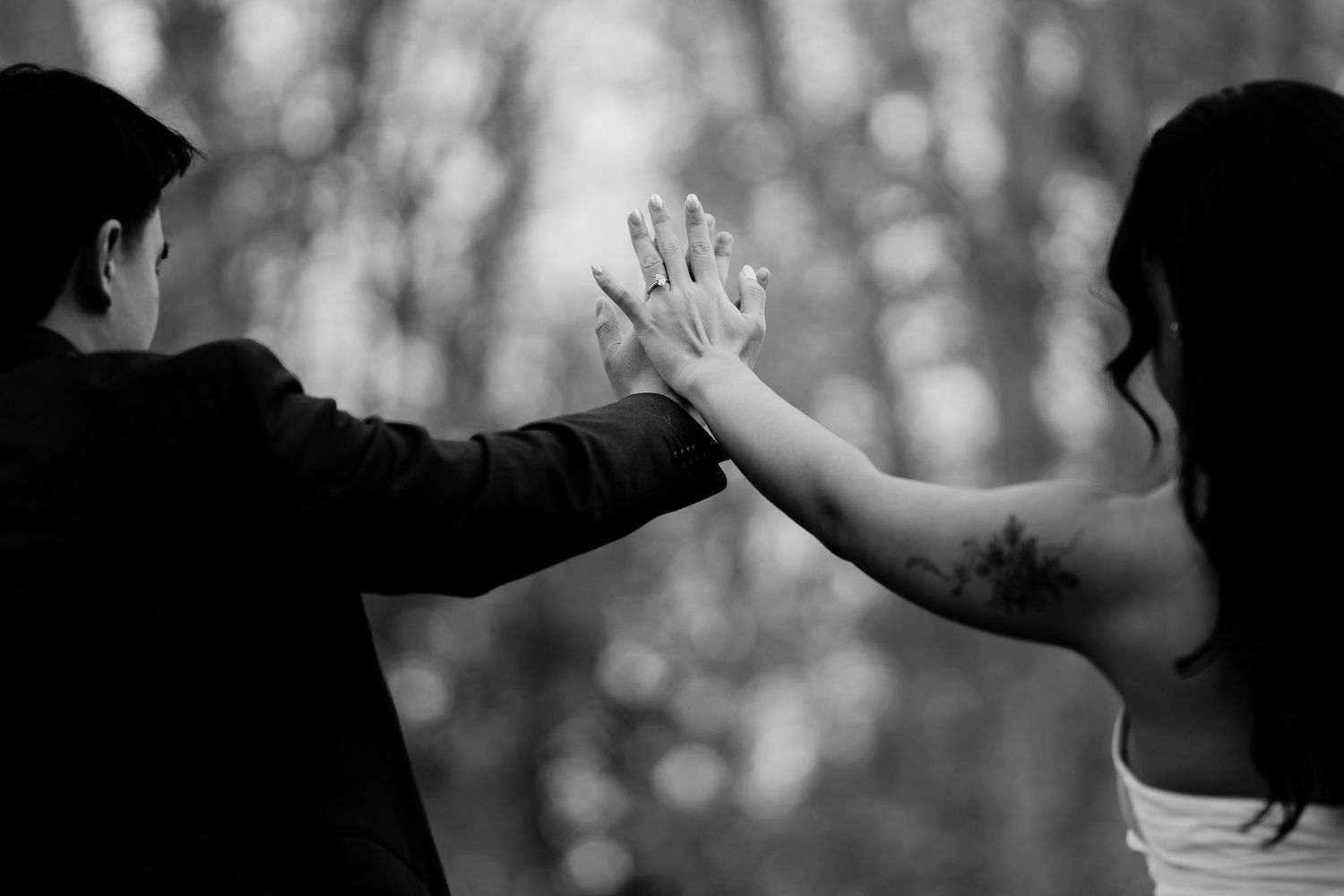 Black and white photo of a couple touching hands, showcasing a wedding ring and a tattoo on the bride's arm.