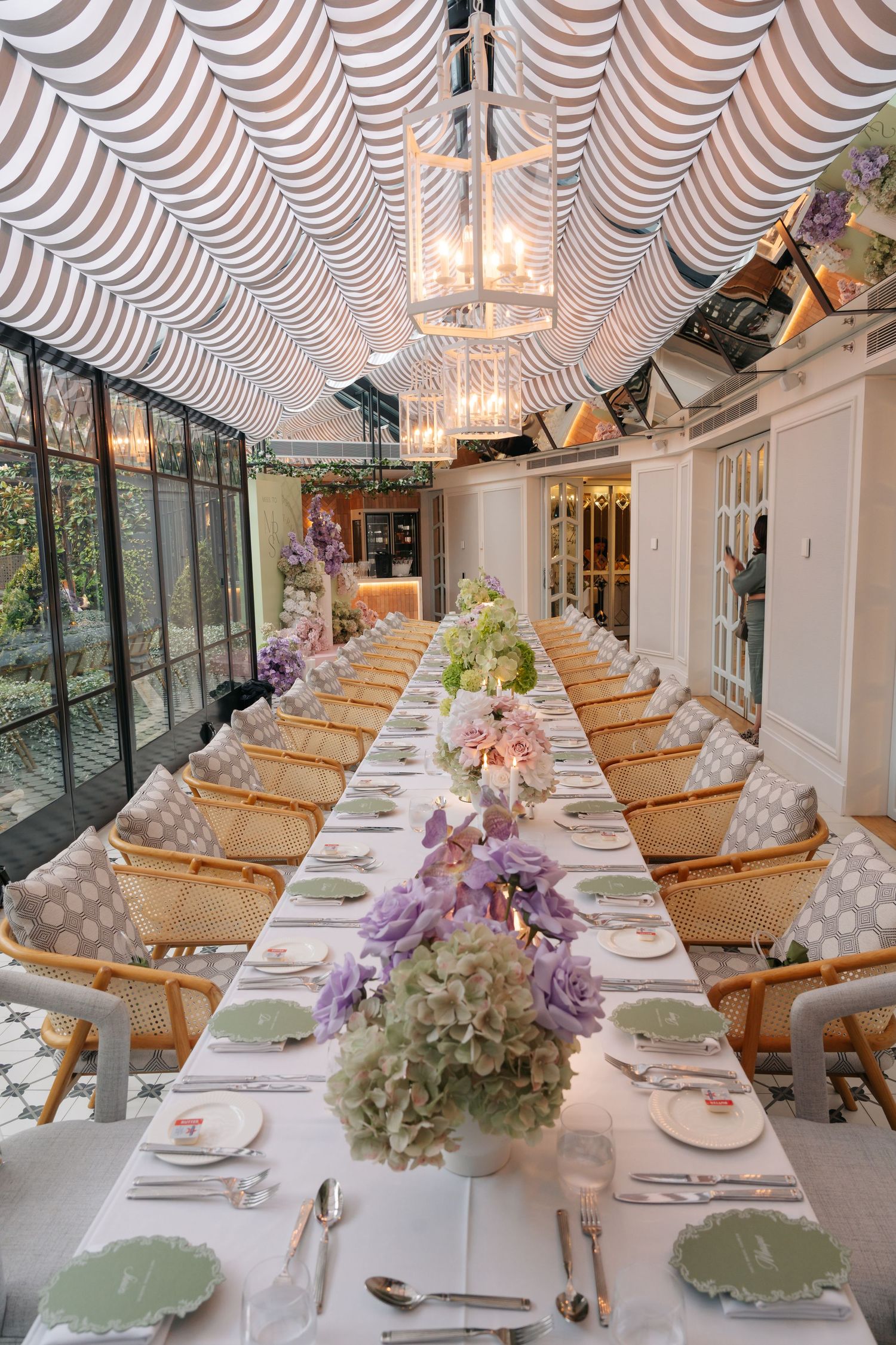 Elegant long dining table with purple hydrangeas, green plates, and striped canopy ceiling in a bright event space.