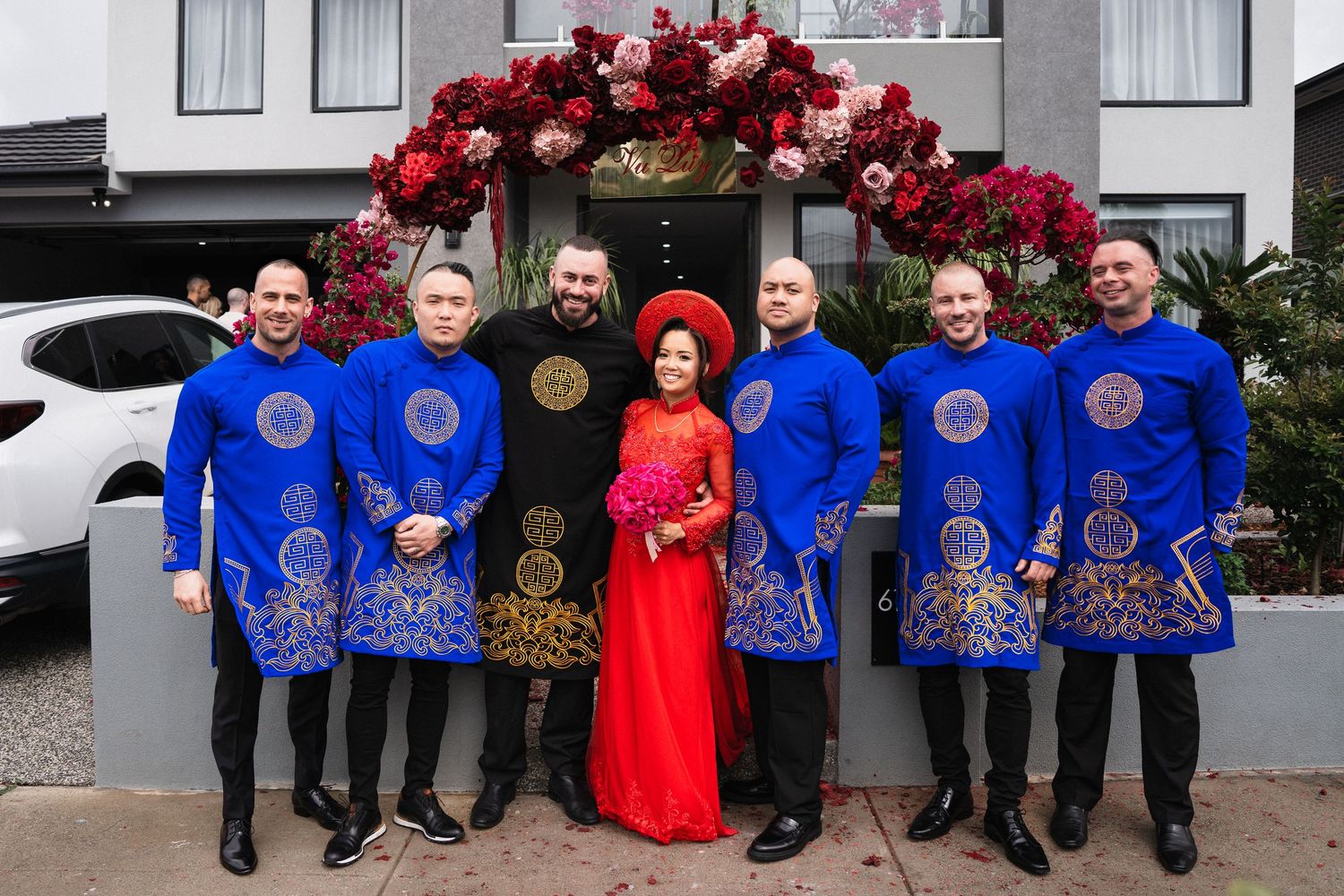 Group of seven men in blue ao dai and couple in red and black traditional Vietnamese wedding attire pose outdoors.