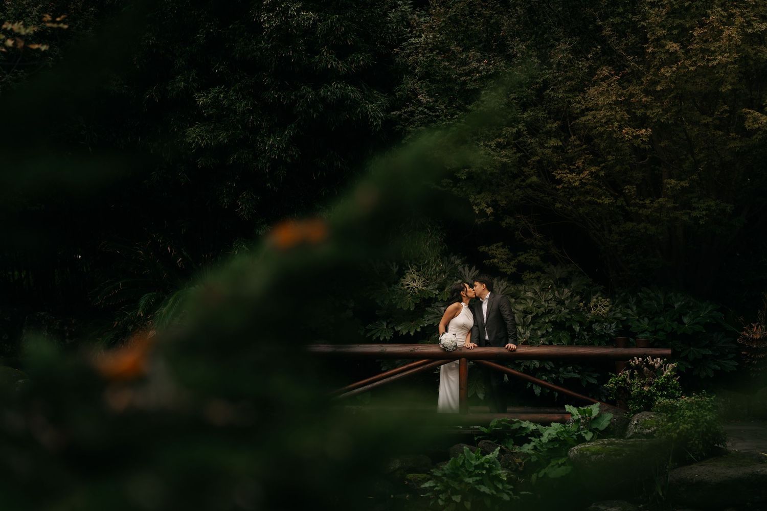 A bride and groom share a romantic moment on a wooden bridge surrounded by lush green forest trees.