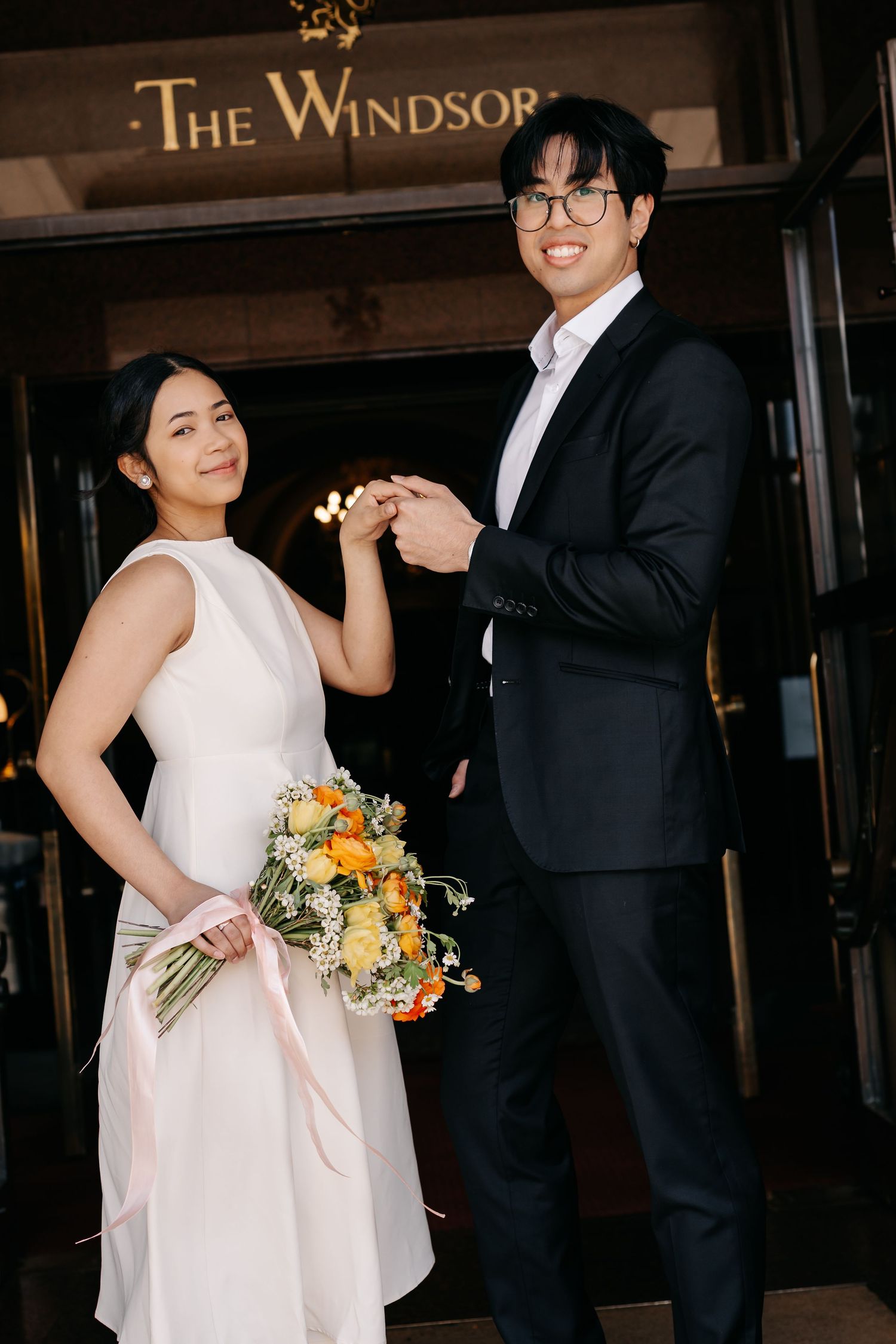 A couple poses playfully outside The Windsor hotel entrance with orange and yellow flowers.