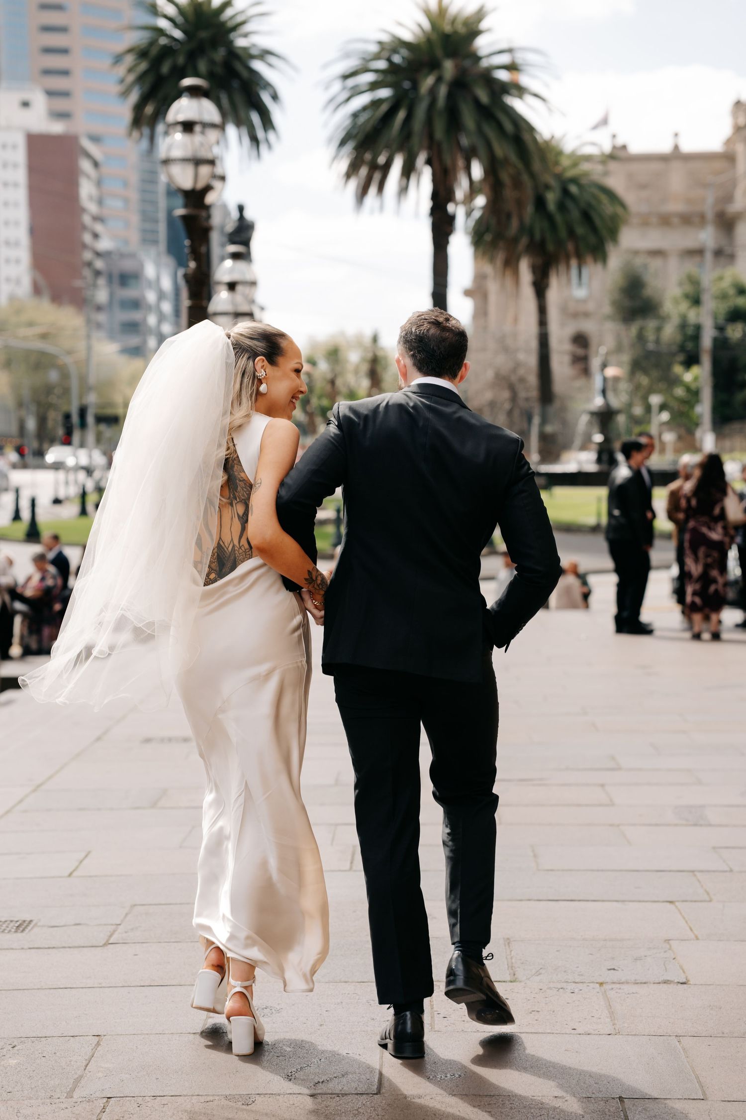 Newlyweds walk together on a palm tree-lined path in an urban setting.