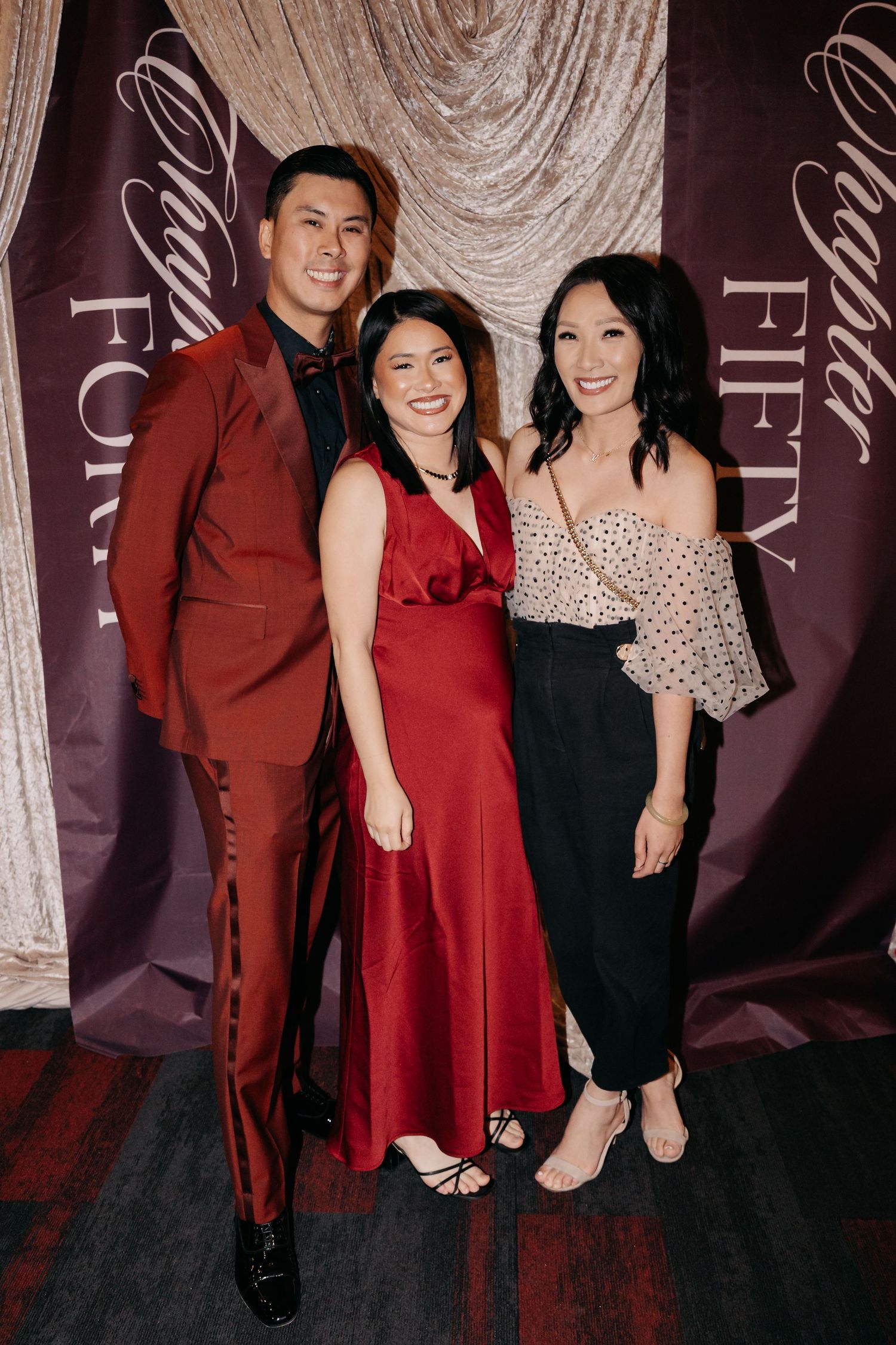 Three elegantly dressed guests pose together at a Chapter Fifty formal event, wearing red and black attire.