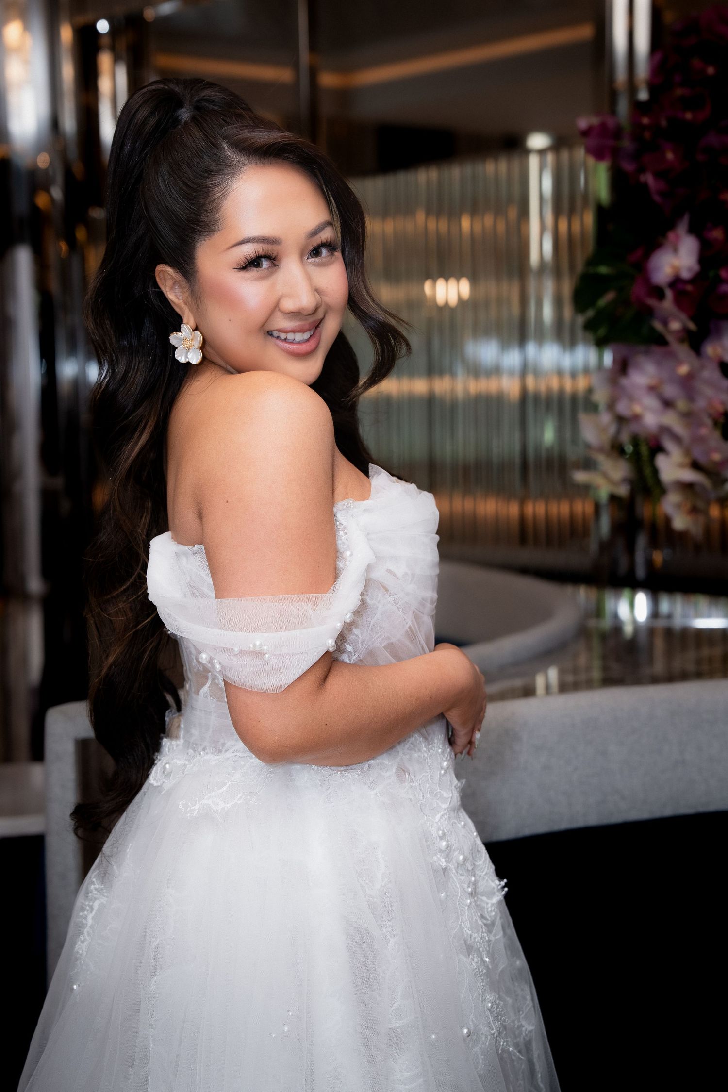 Smiling bride in white off-shoulder wedding gown with floral earrings poses elegantly in a luxurious venue.