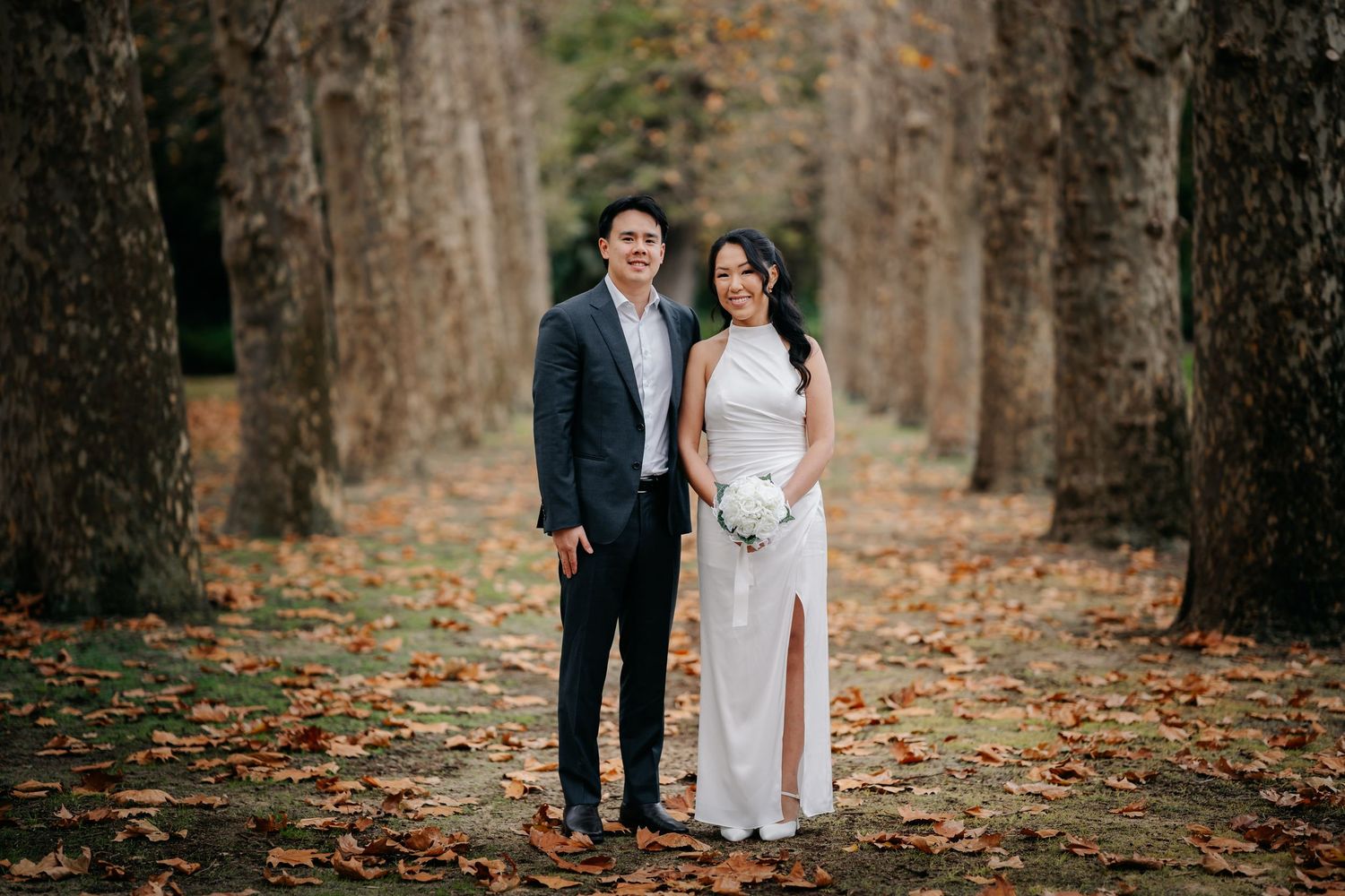 Couple in wedding attire posing among autumn trees with fallen leaves on ground.