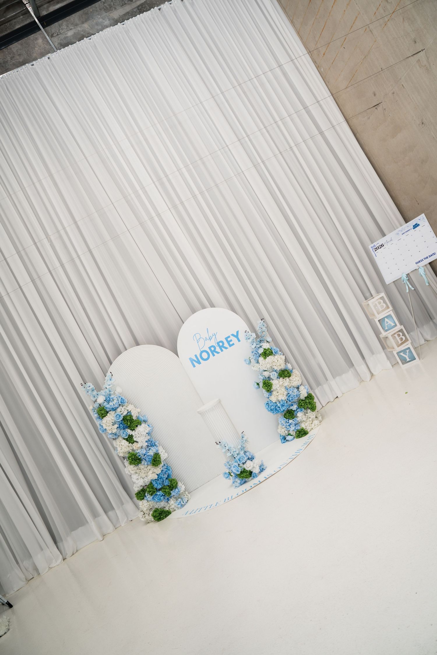 White heart-shaped baby shower backdrop with blue hydrangeas and Baby Norrey sign against draped white curtain.