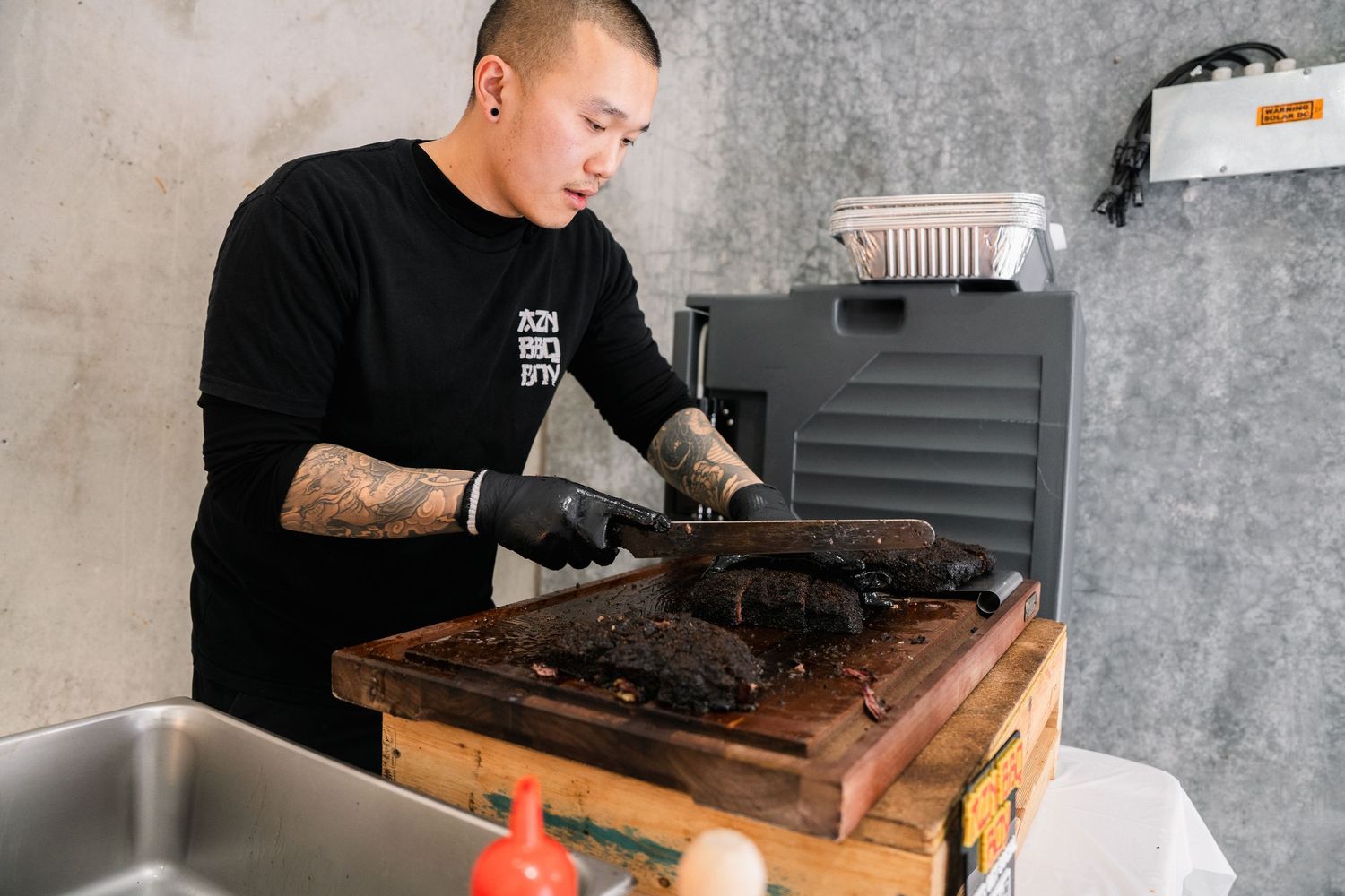 Tattooed chef in black gloves slices smoked brisket on wooden cutting board in modern kitchen.