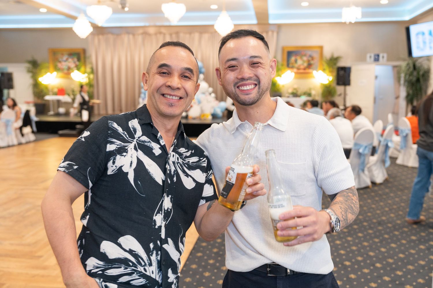 Two smiling men pose together at a banquet hall event, one holding a beer bottle and glass.