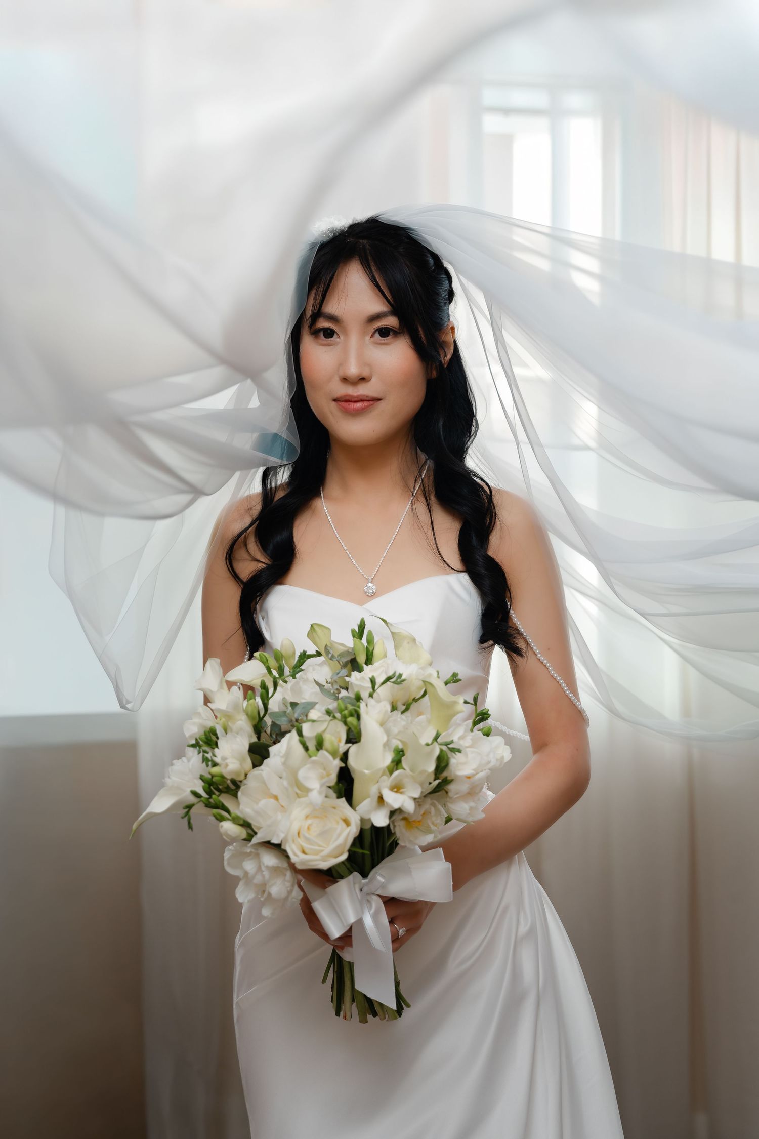 Elegant Asian bride in white satin gown holds white floral bouquet with veil flowing behind her.