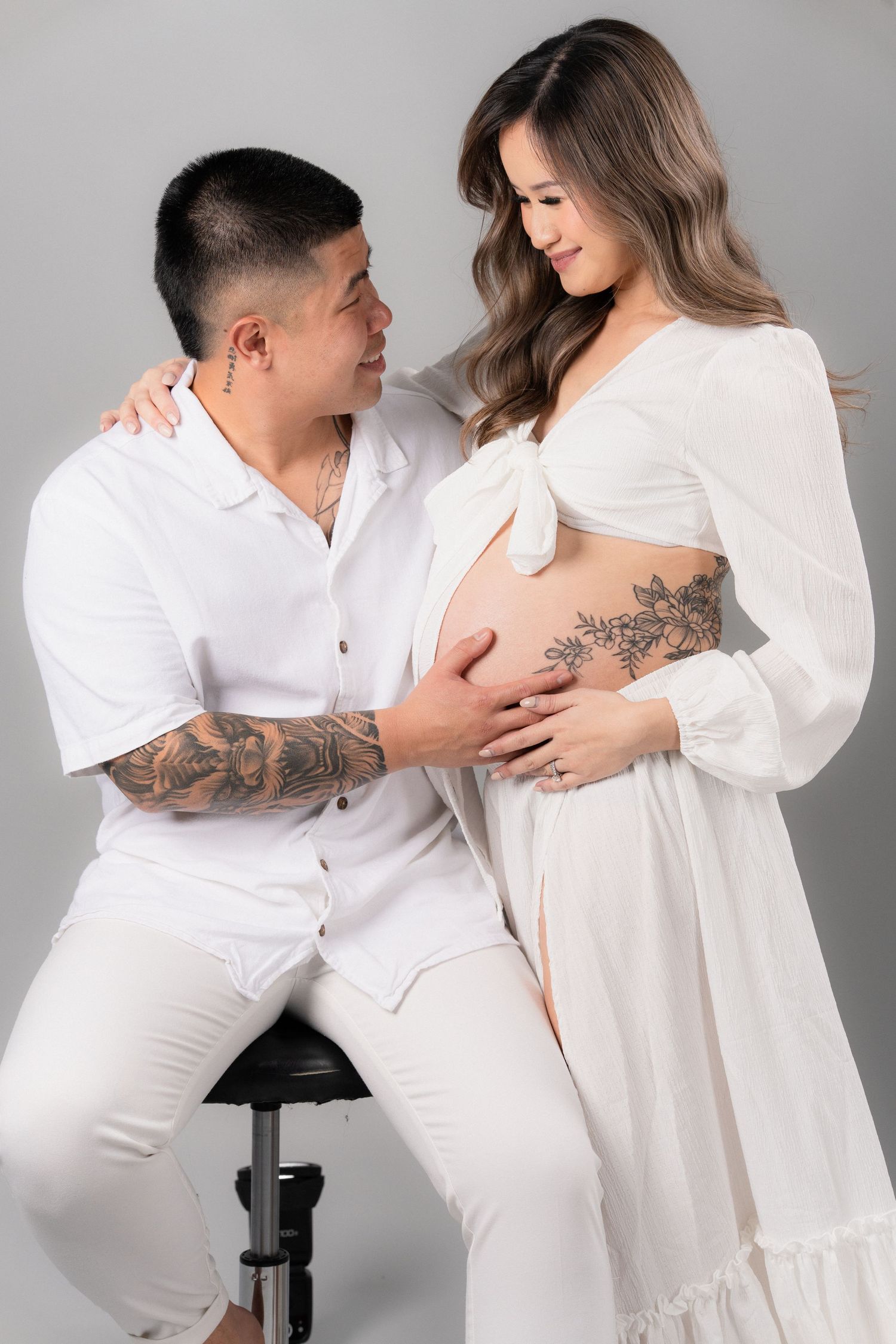 Couple in white outfits posing for maternity photo, man sitting on stool touching pregnant woman's belly.