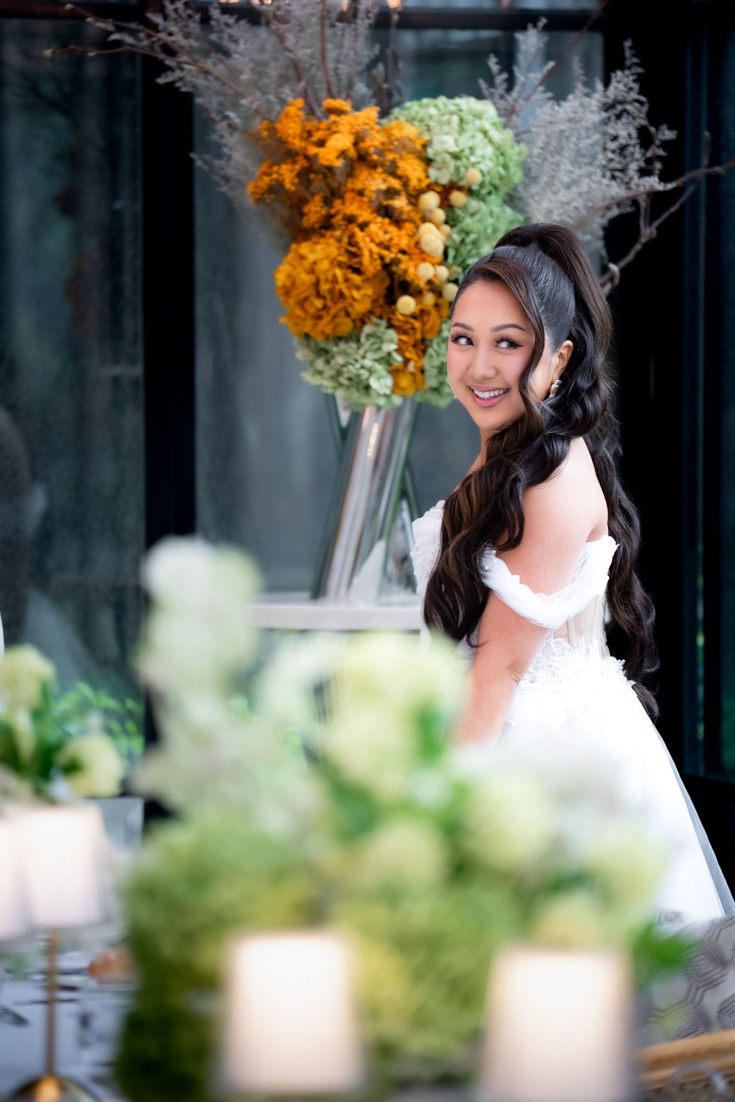 Smiling bride in white off-shoulder gown poses at elegantly decorated table with lush floral arrangements.