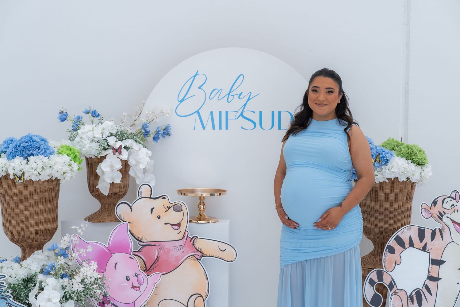Pregnant woman in blue dress at Baby Shower with Winnie the Pooh decorations and floral backdrop.