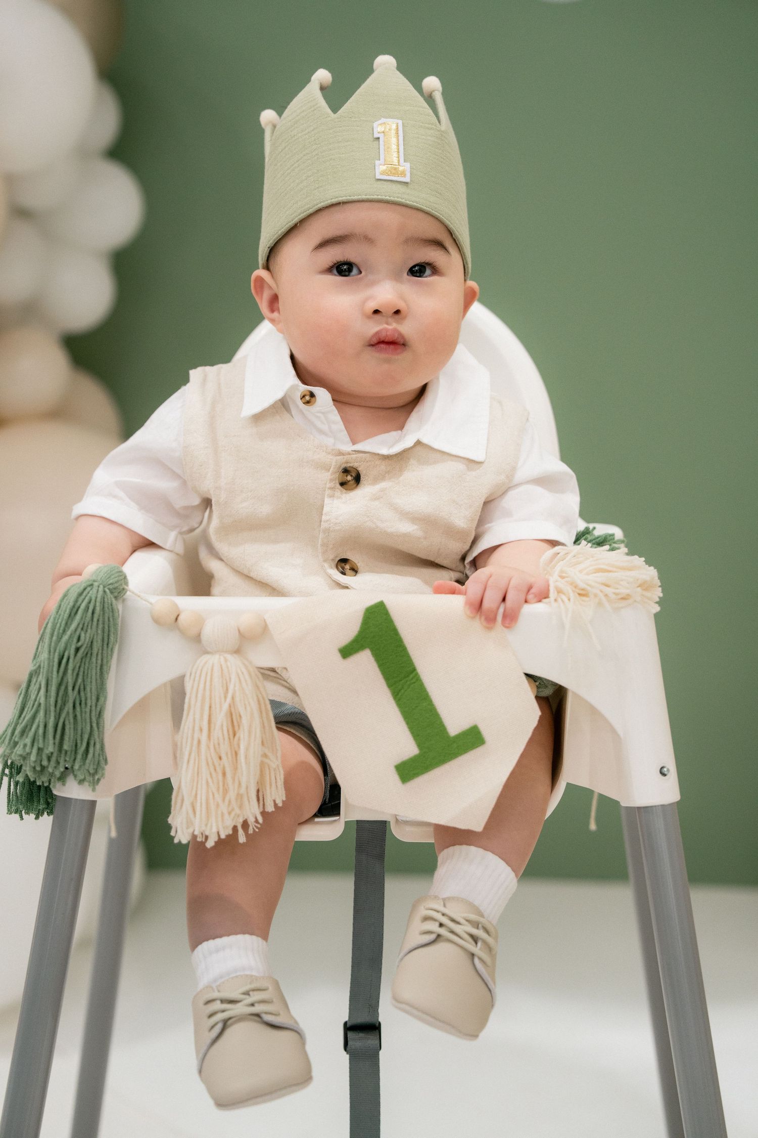 Cute baby boy in cream outfit and green number one crown sits in high chair at first birthday party.