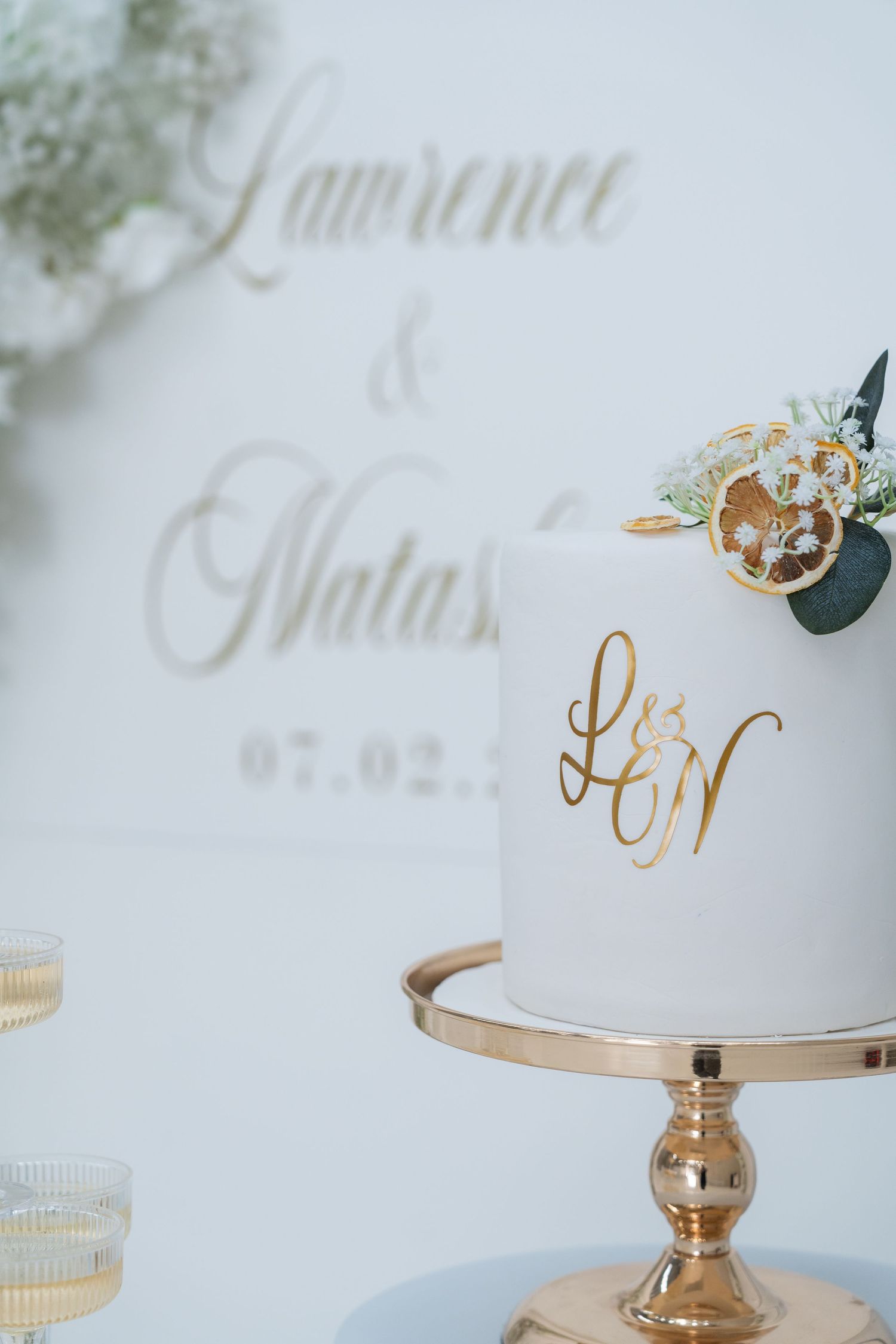 Elegant white wedding cake with gold L&N monogram on a gold stand, decorated with flowers, for Lawrence and Natasha.