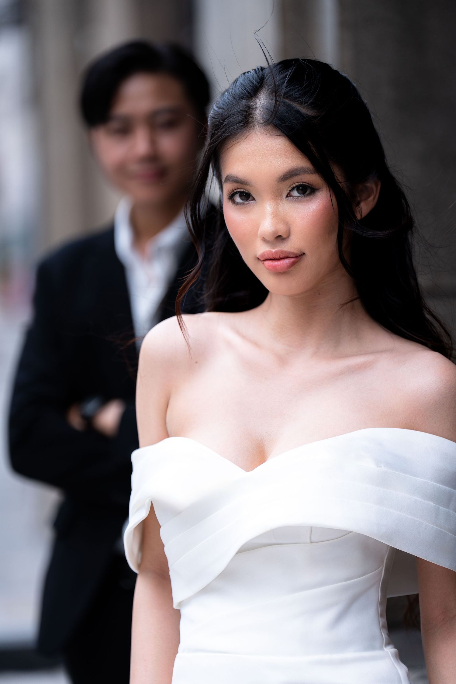 Elegant bride in off-shoulder white wedding gown with natural makeup and flowing dark hair.