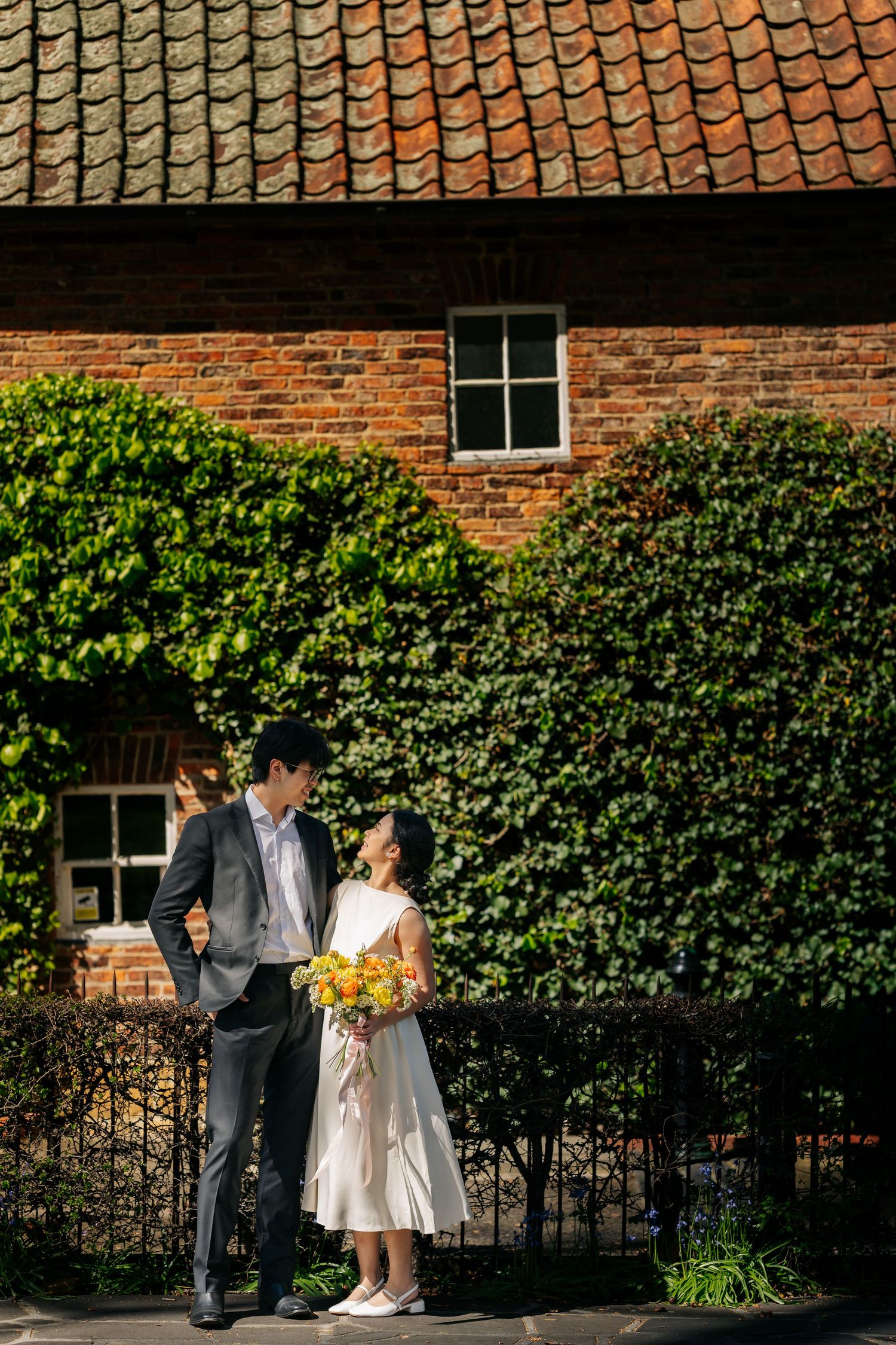 Couple walks together along ivy-covered brick wall in formal wedding attire.