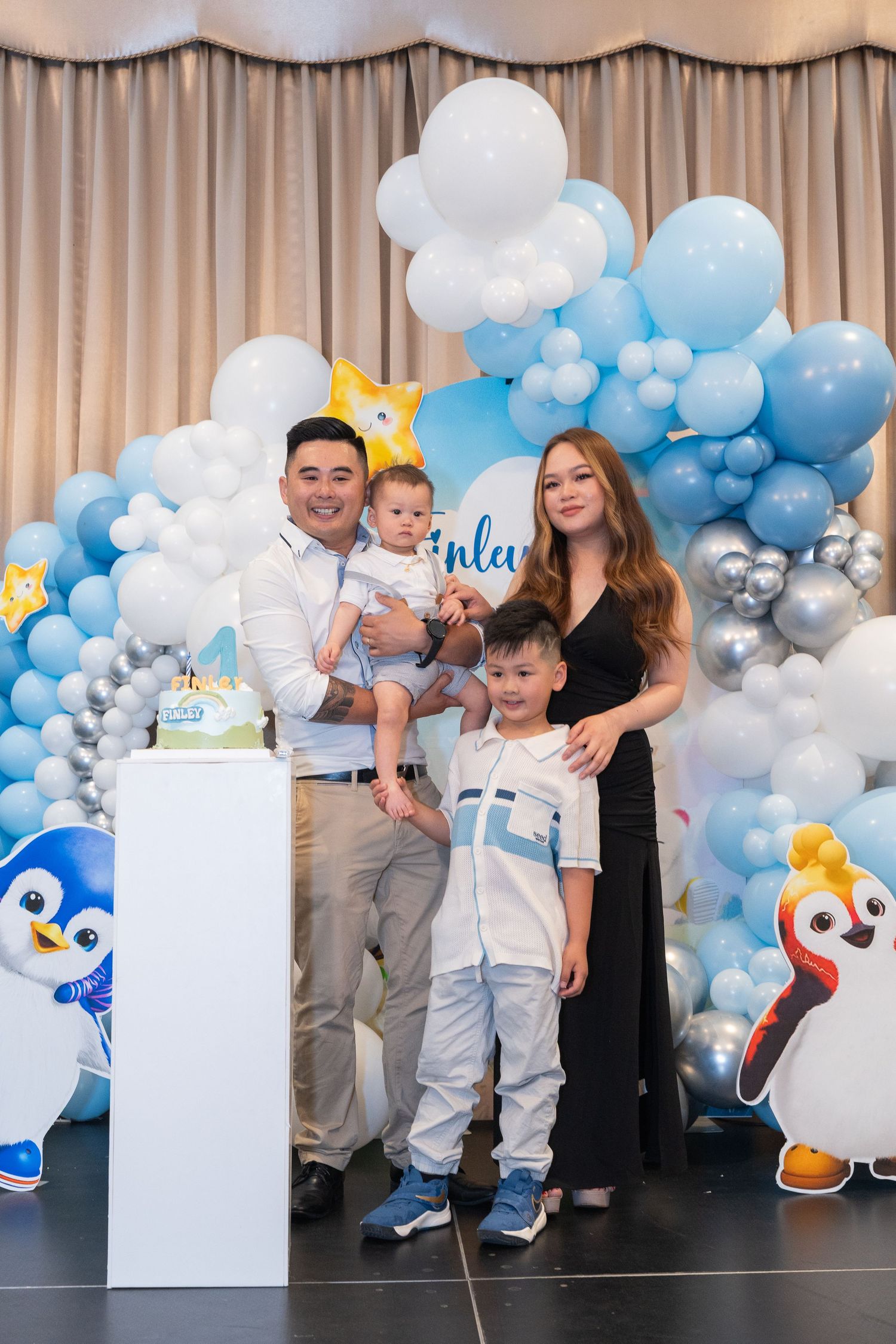 Family posing at a baby's first birthday party with blue balloon arch, penguin decorations, and birthday cake.