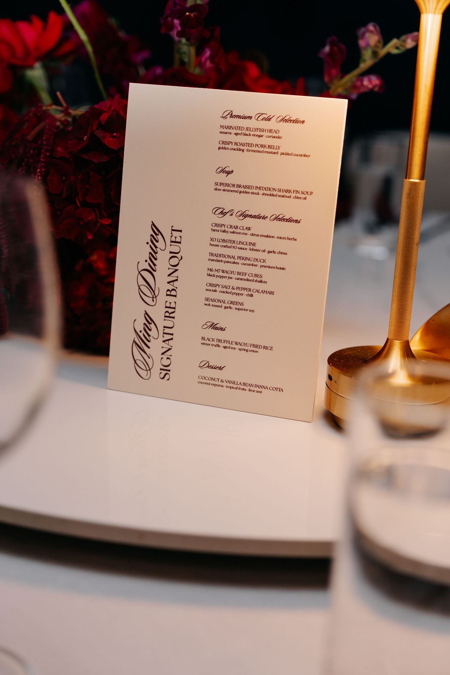 Elegant fine dining banquet menu card displayed on a white plate setting with gold candlestick and red floral arrangement.