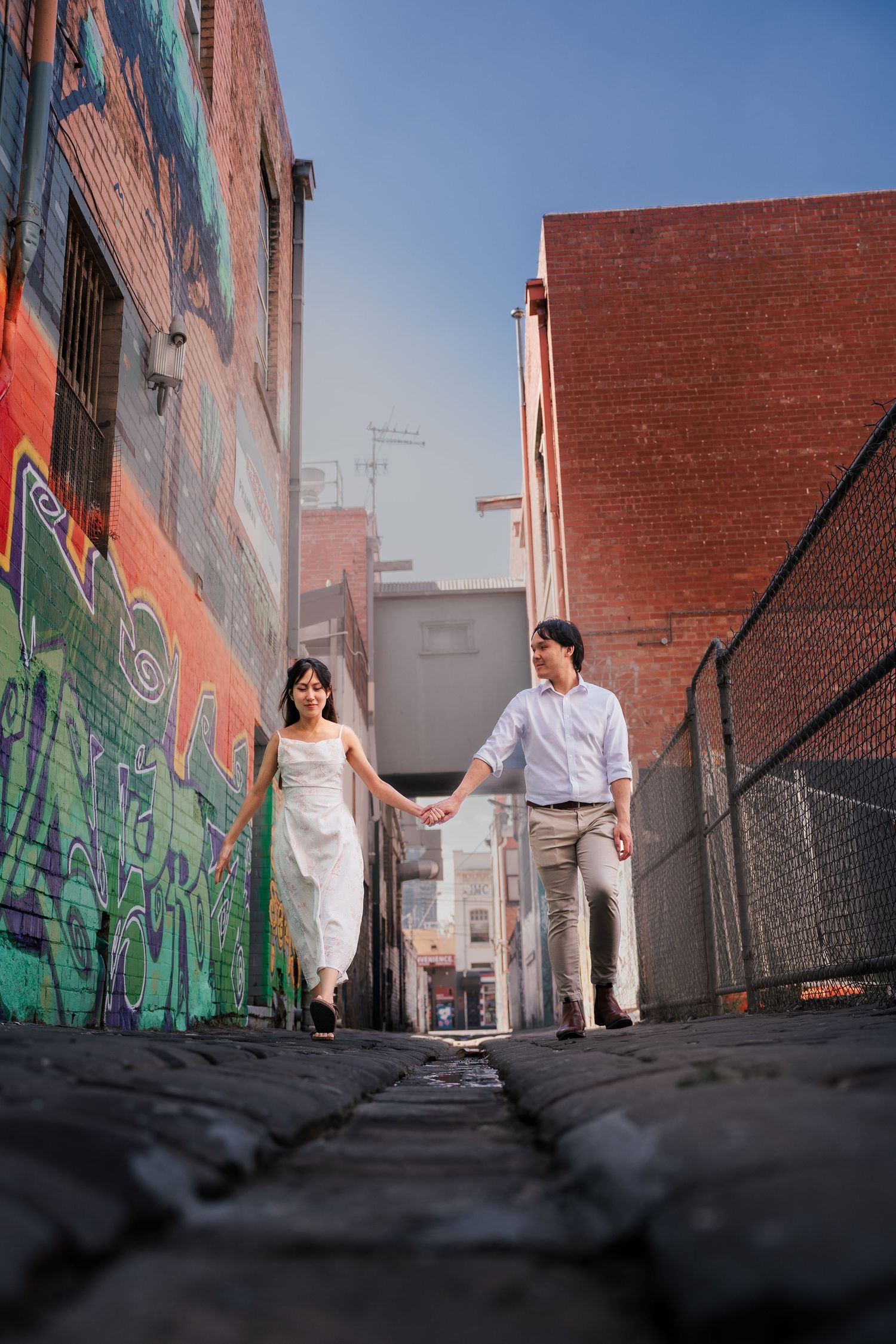 Couple holding hands walking through a colorful graffiti alley with brick buildings under a clear blue sky.