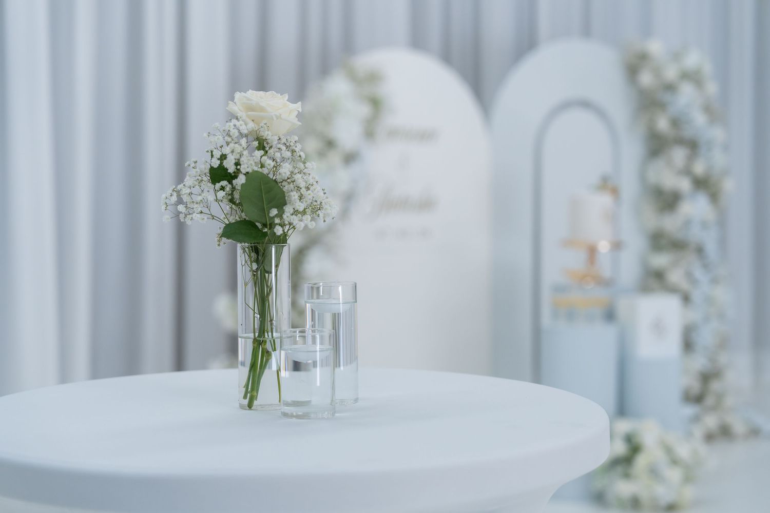 Elegant wedding table with white roses in glass vase, soft blue backdrop with floral arch decor.