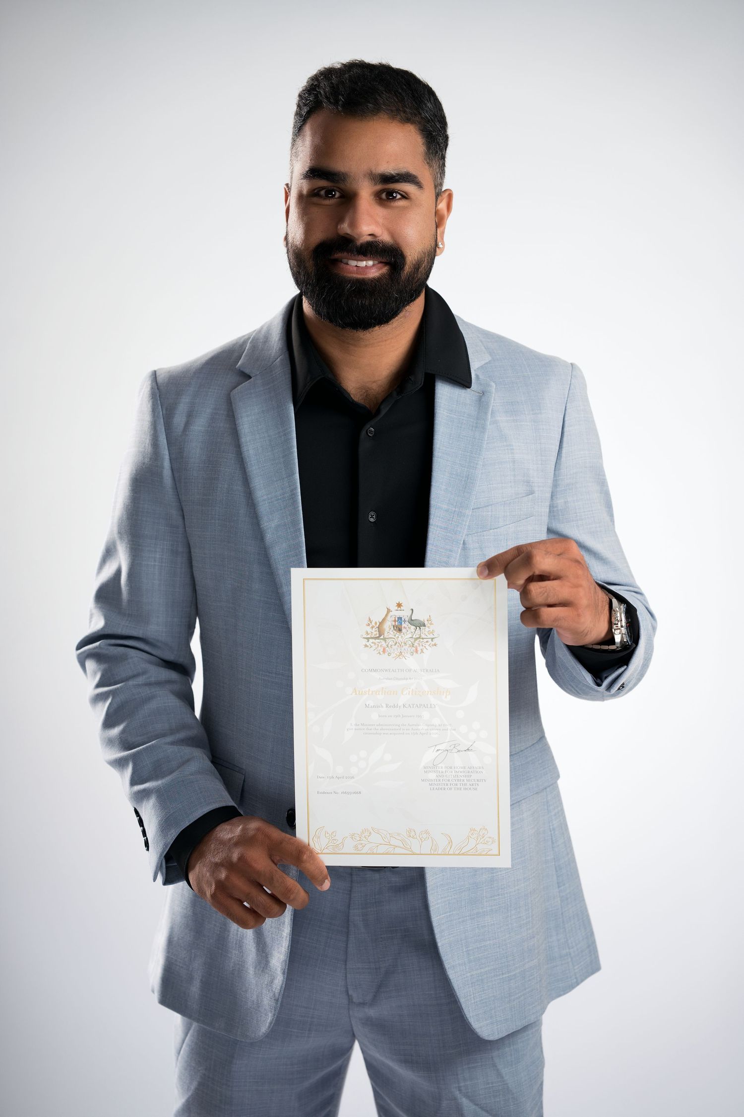 Smiling bearded man in light blue suit proudly holding an Australian citizenship certificate against white background.