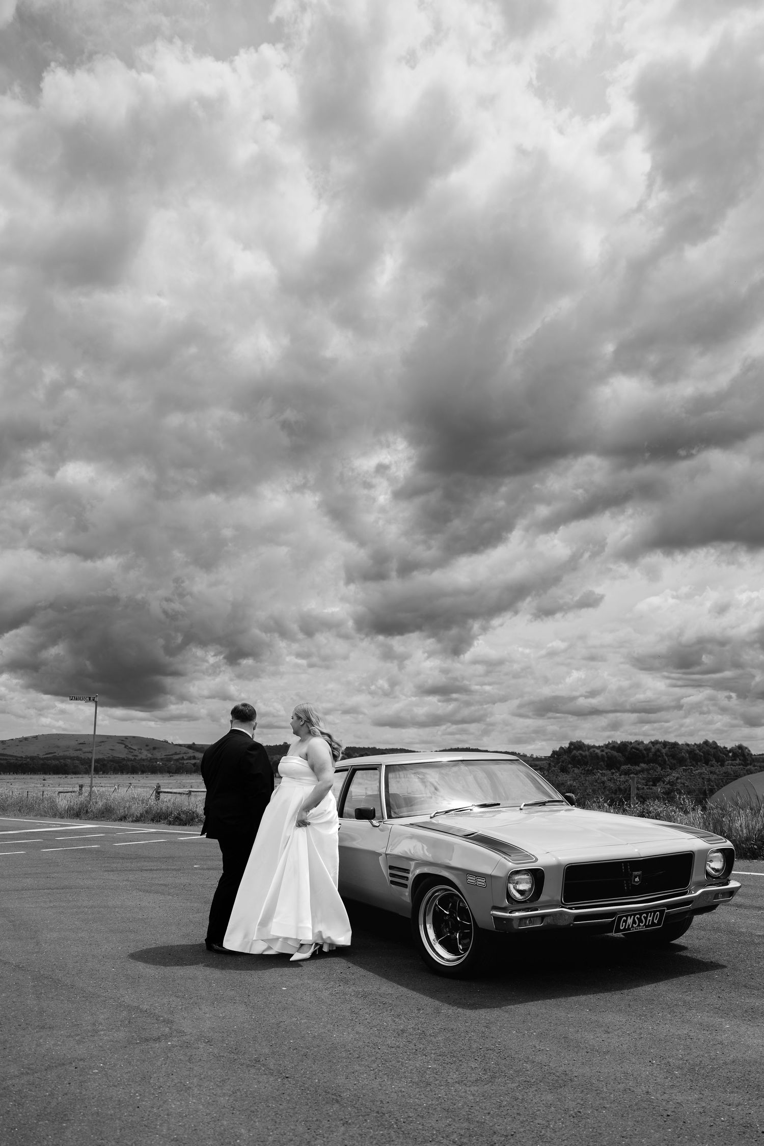 Bride and groom pose beside a classic muscle car under dramatic cloudy skies in a black and white wedding photo.