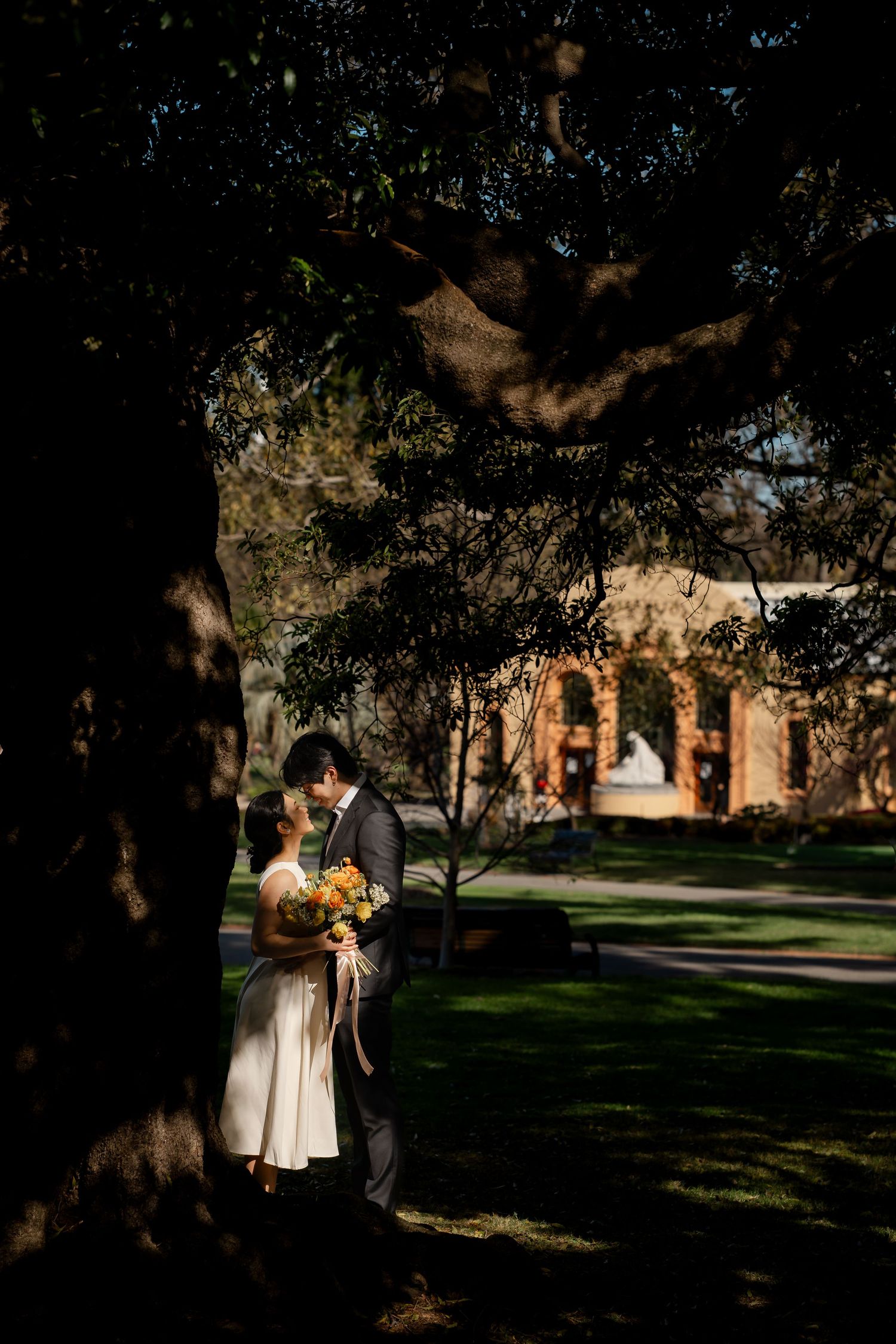 A couple stands in dappled sunlight among trees in a romantic garden setting.